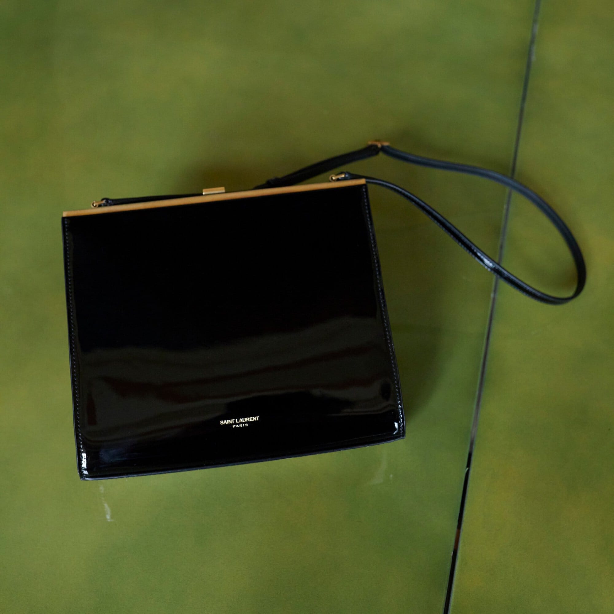 Elevated view of a black patent leather handbag with a gold frame and strap handle on top of a green and brown marbled surface.