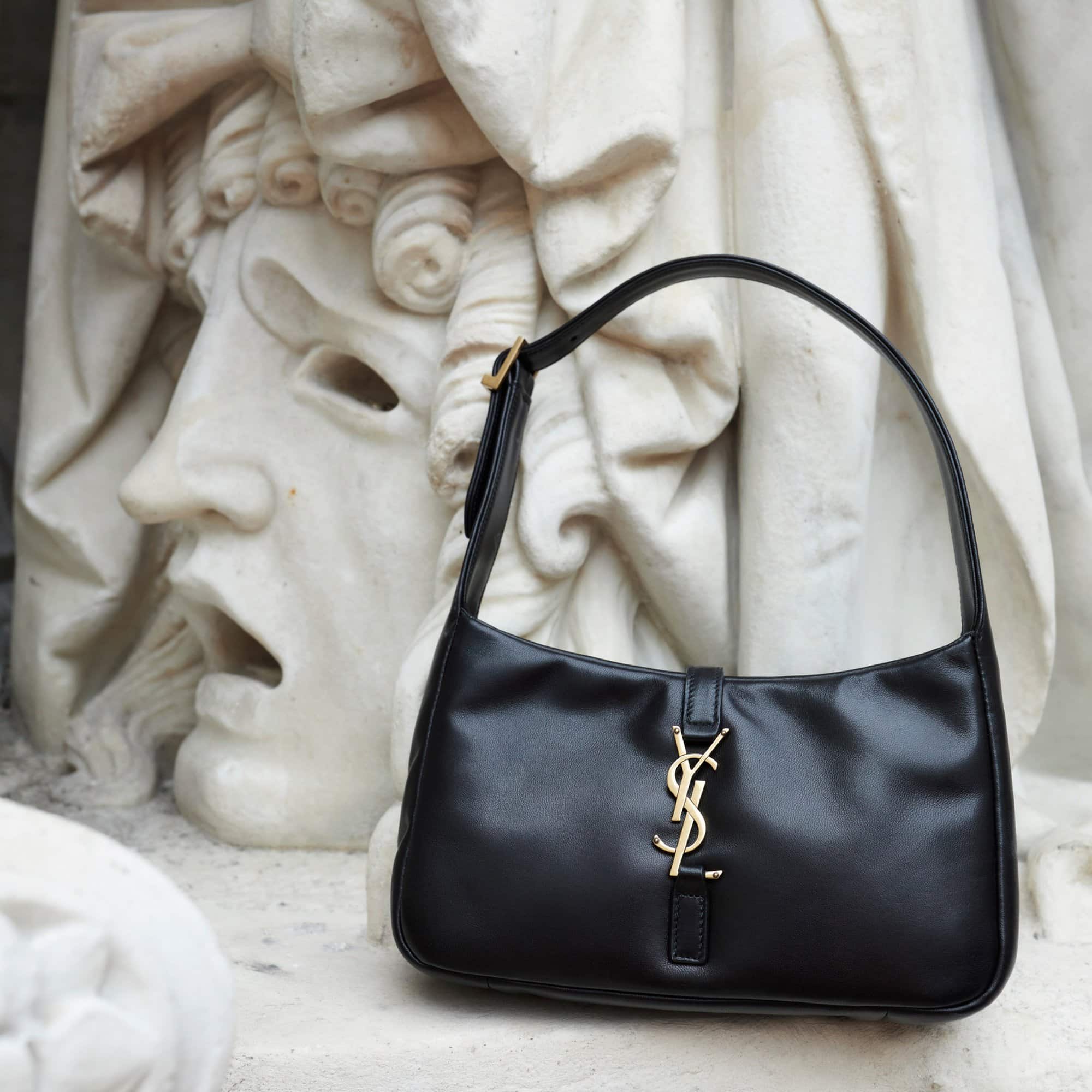 Black Saint Laurent purse with gold logo hardware, displayed on a weathered stone statue with head and curly hair details, medium shot.