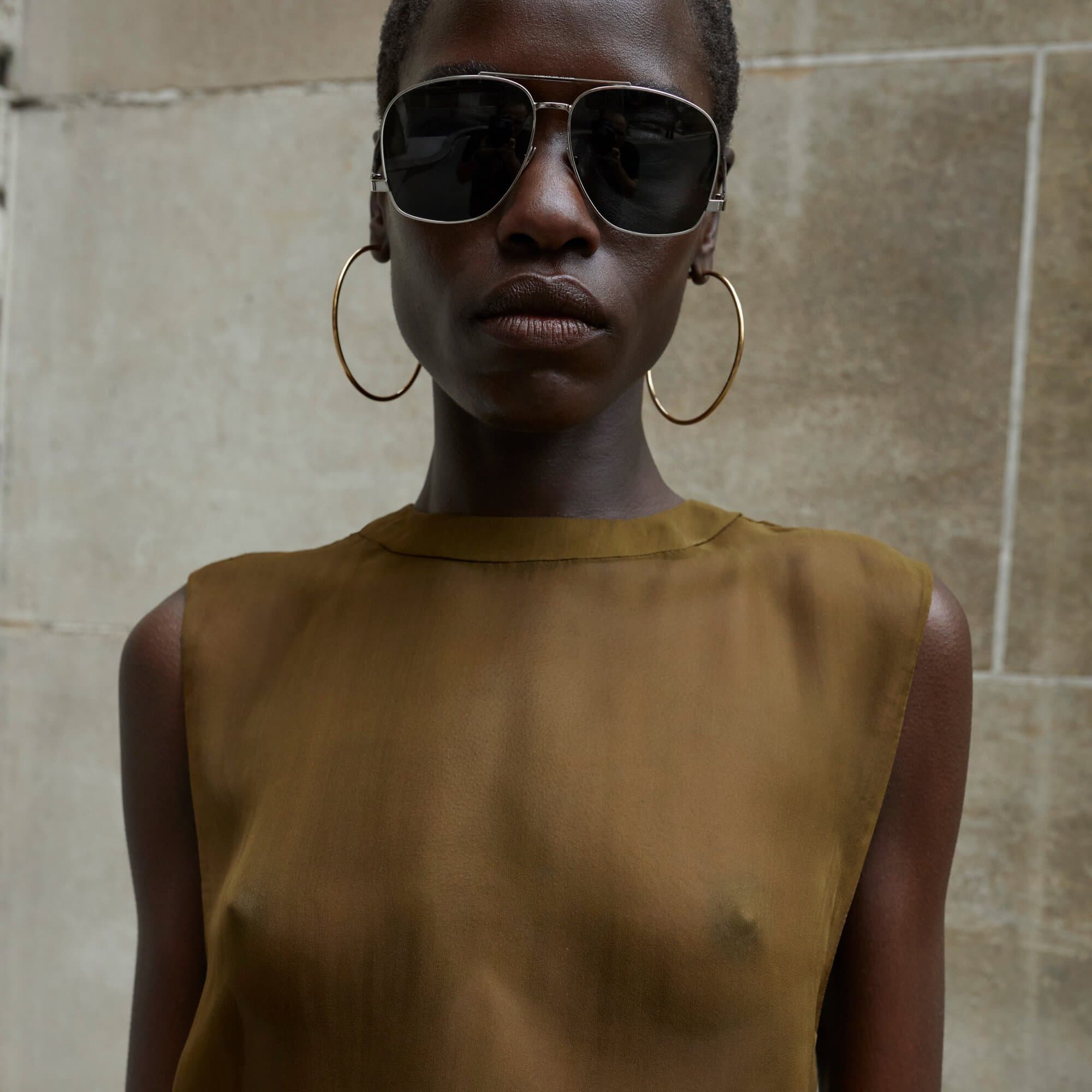 Close-up of a stylish dark-skinned woman with sunglasses and hoop earrings wearing an olive-green top posing against a neutral wall.