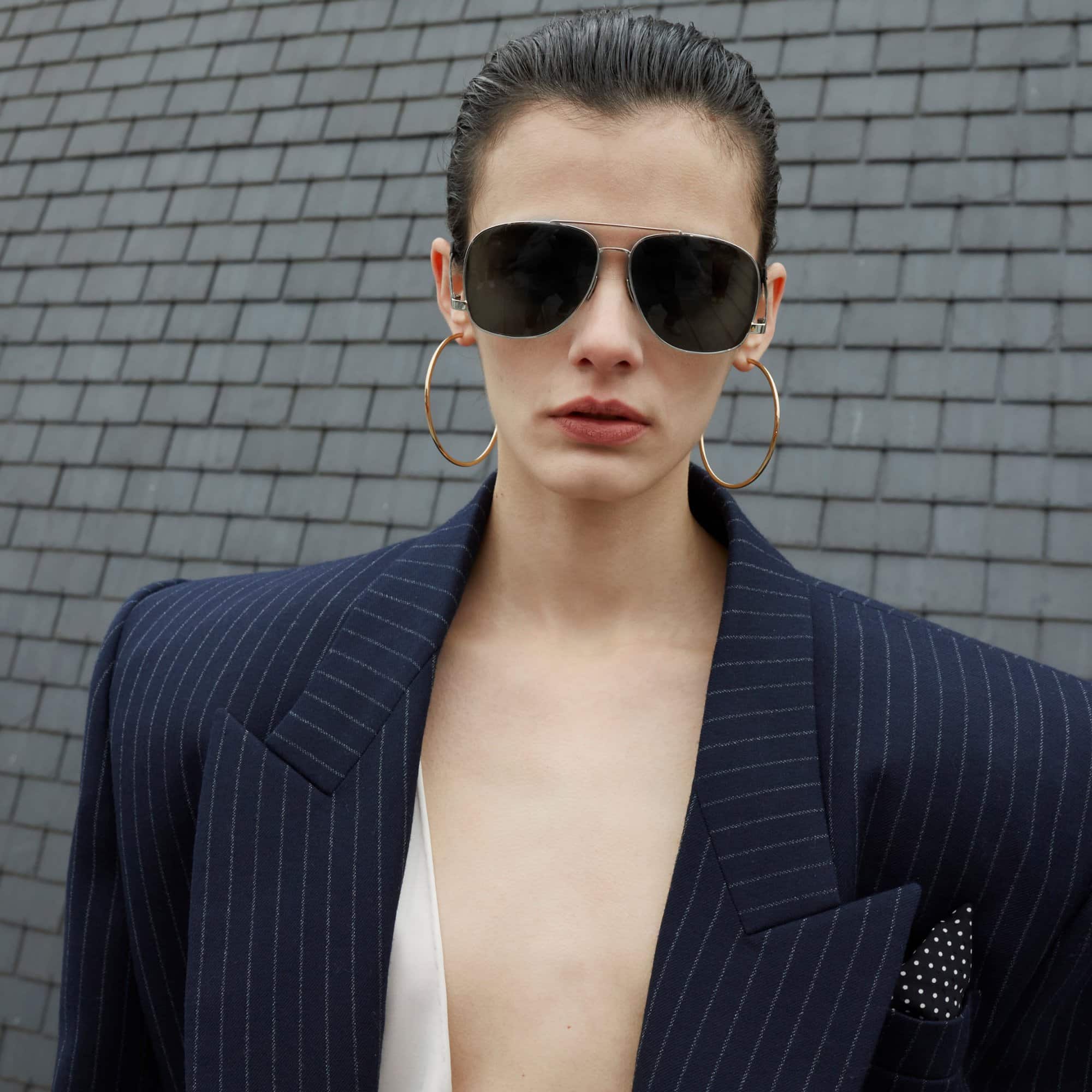 Woman poses in a pinstripe blazer with aviator sunglasses and gold hoop earrings against a gray brick wall in a medium studio shot.