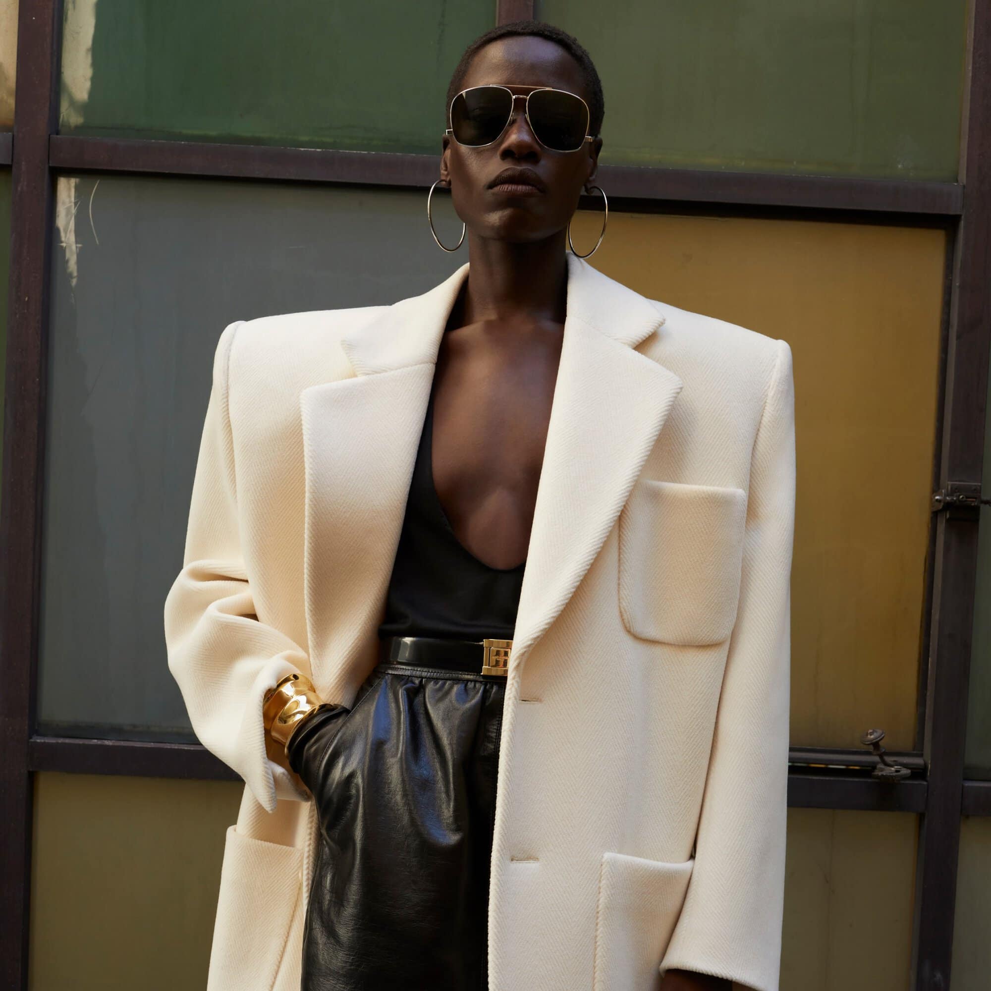 Fashionable dark-skinned woman in sunglasses and a blazer poses in front of a colorful window wearing leather skirt and gold jewelry.