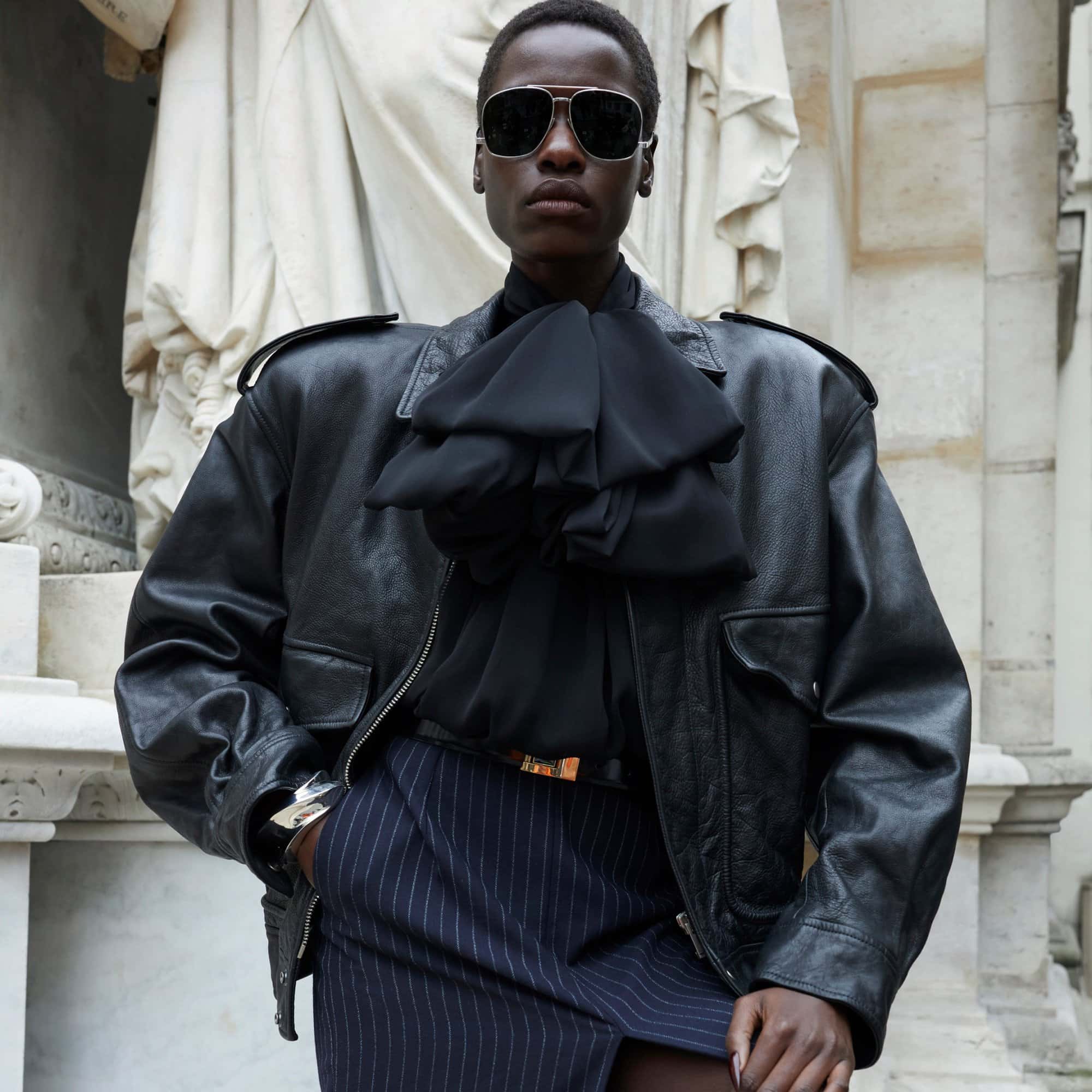 Woman in aviator sunglasses poses outdoors wearing a black leather bomber jacket, ruffled blouse, and pinstripe skirt against a stone wall.