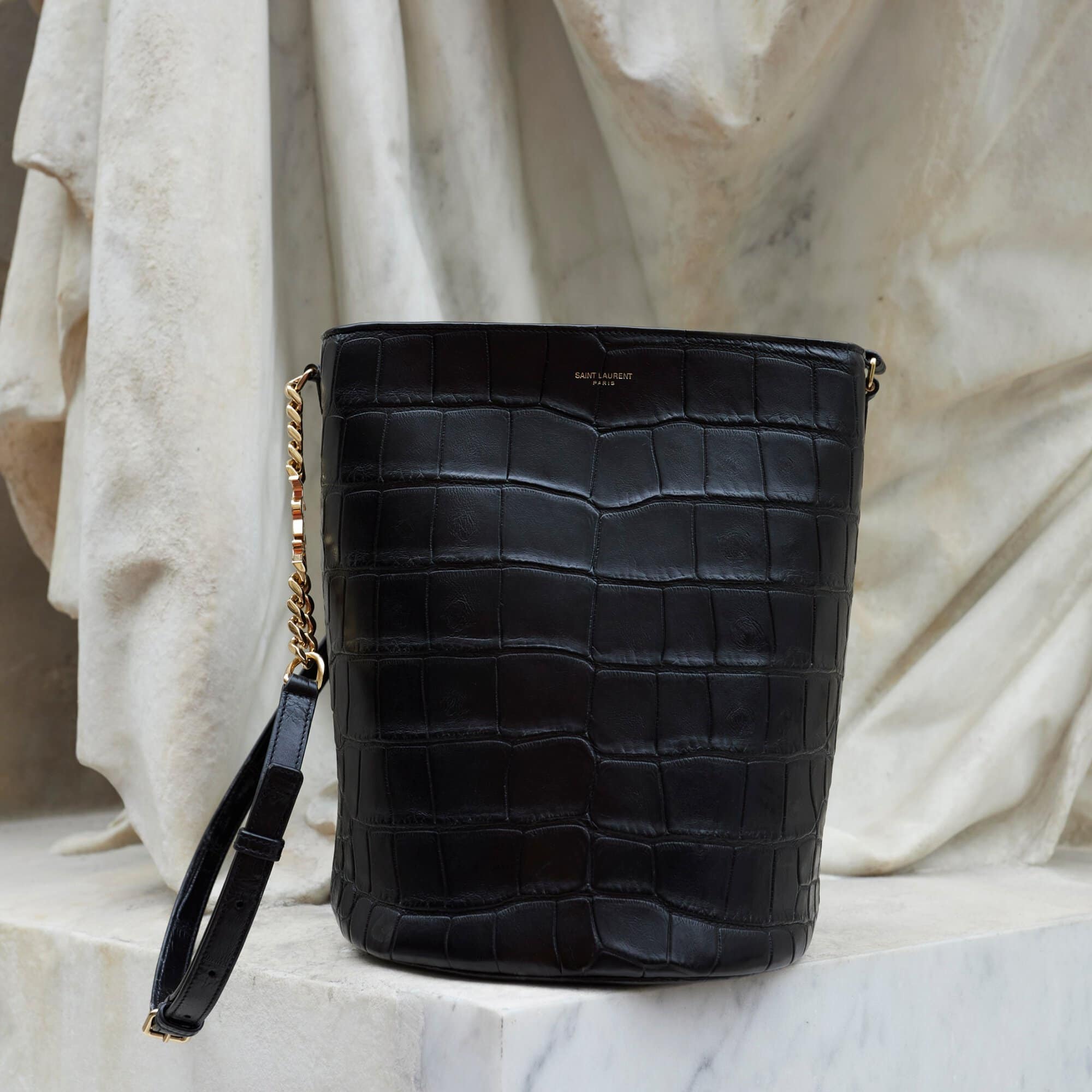 Black Saint Laurent bucket bag with gold chain detail displayed indoors against a white marble sculpture. Fashion product shot.