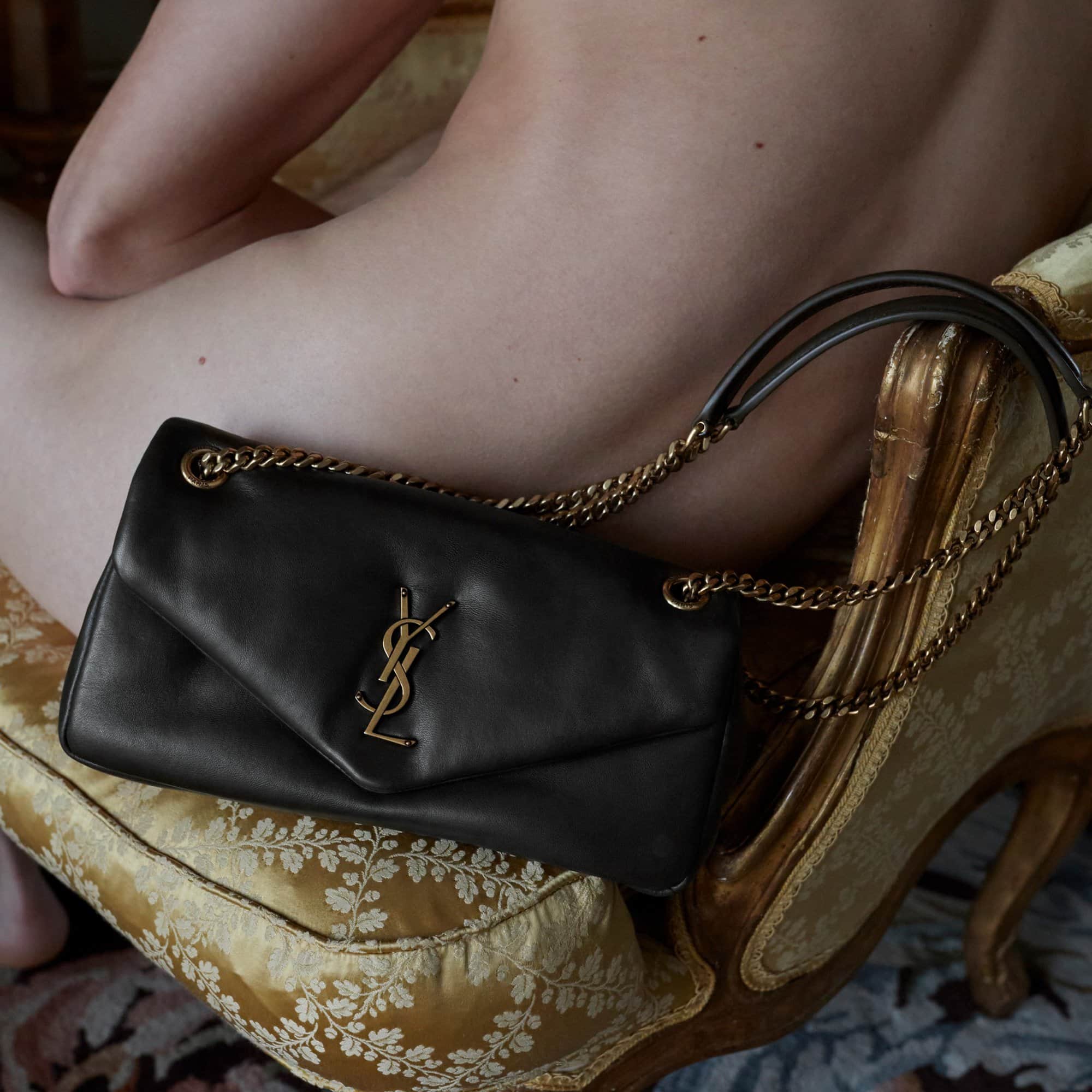 Close-up of a black Saint Laurent shoulder bag with gold chain straps and logo on a gold patterned cushion next to a person.