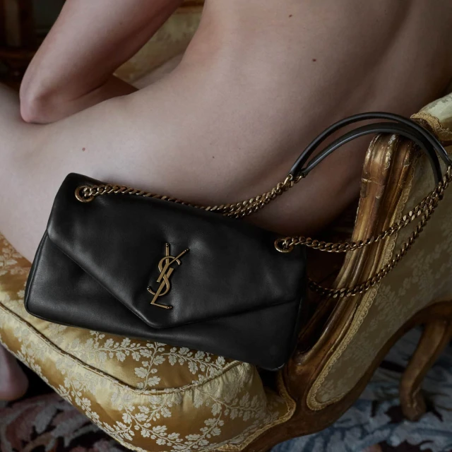 Close-up of a black Saint Laurent shoulder bag with gold chain straps and logo on a gold patterned cushion next to a person.