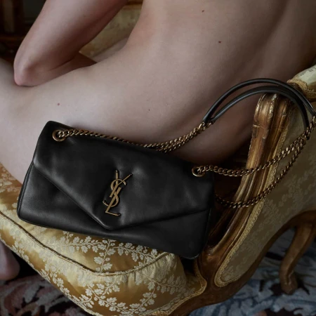 Close-up of a black Saint Laurent shoulder bag with gold chain straps and logo on a gold patterned cushion next to a person.
