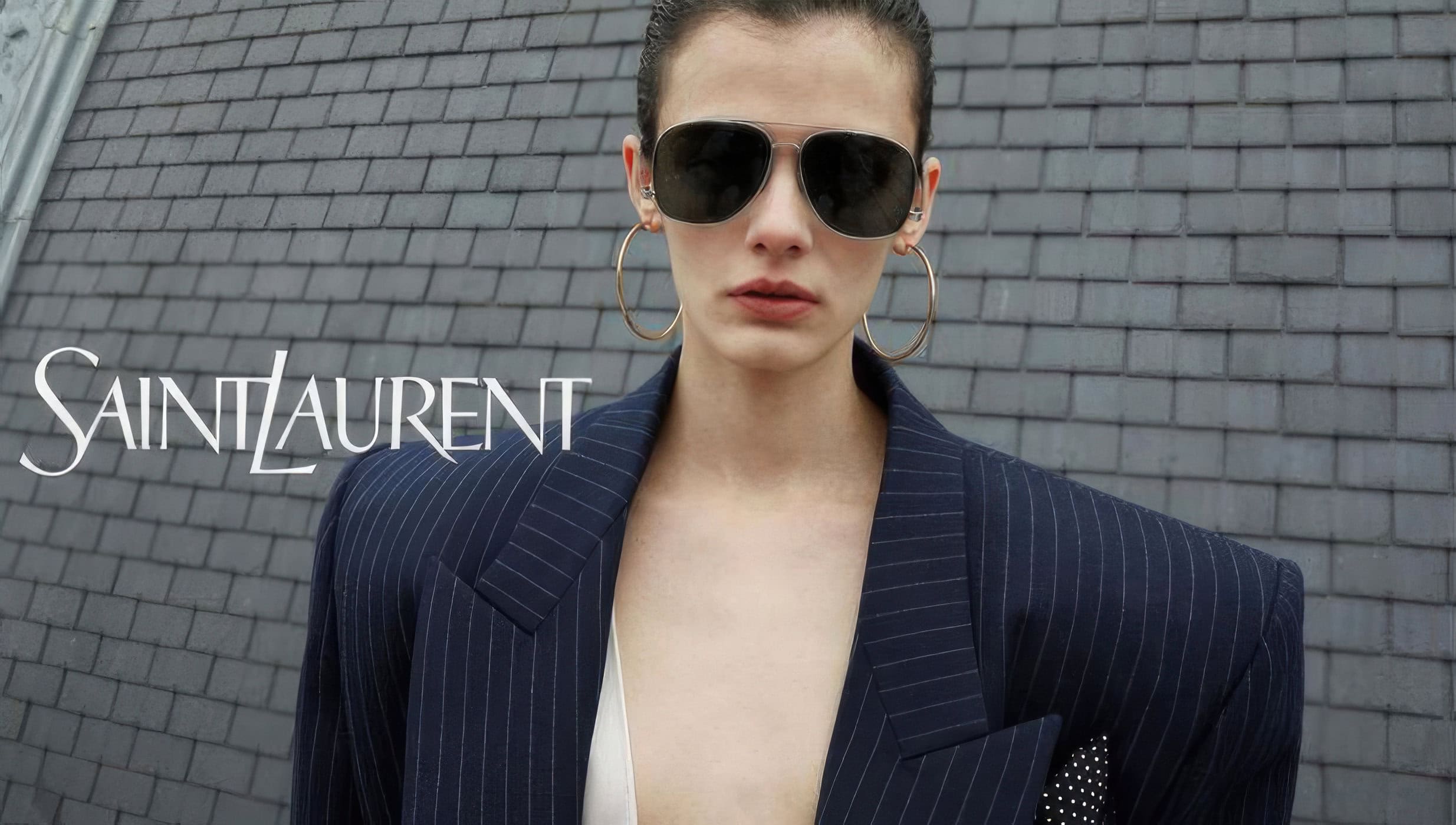 Fashion shot featuring a woman wearing sunglasses and a pinstripe blazer with Saint Laurent logo against a grey shingled roof.
