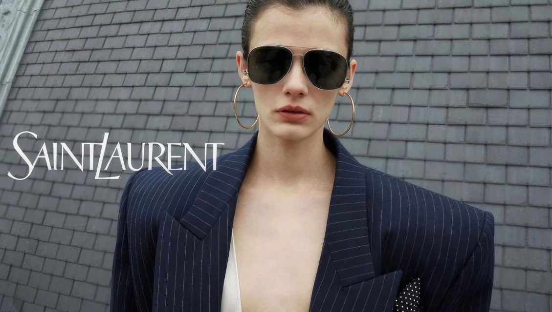 Fashion shot featuring a woman wearing sunglasses and a pinstripe blazer with Saint Laurent logo against a grey shingled roof.