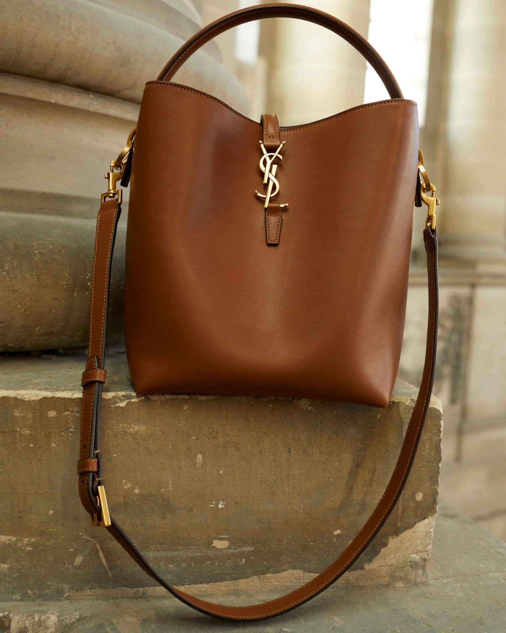 Brown leather handbag with a handle and long strap, featuring a gold-colored logo, resting on stone steps in an indoor setting.