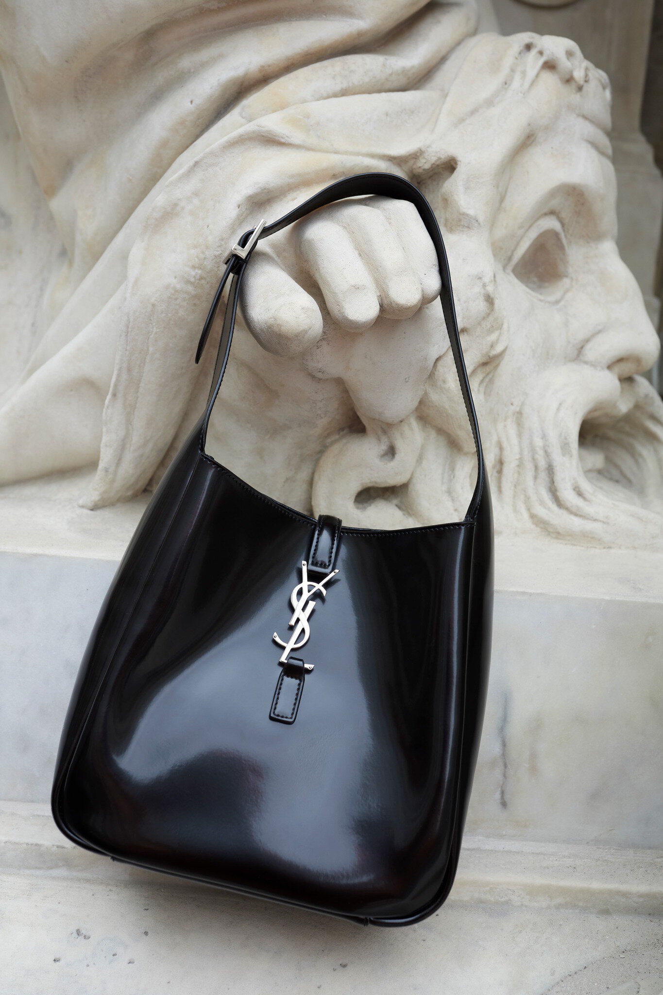 Black Saint Laurent hobo bag with silver YSL logo detail in front of a stone sculpture, luxury fashion photography.