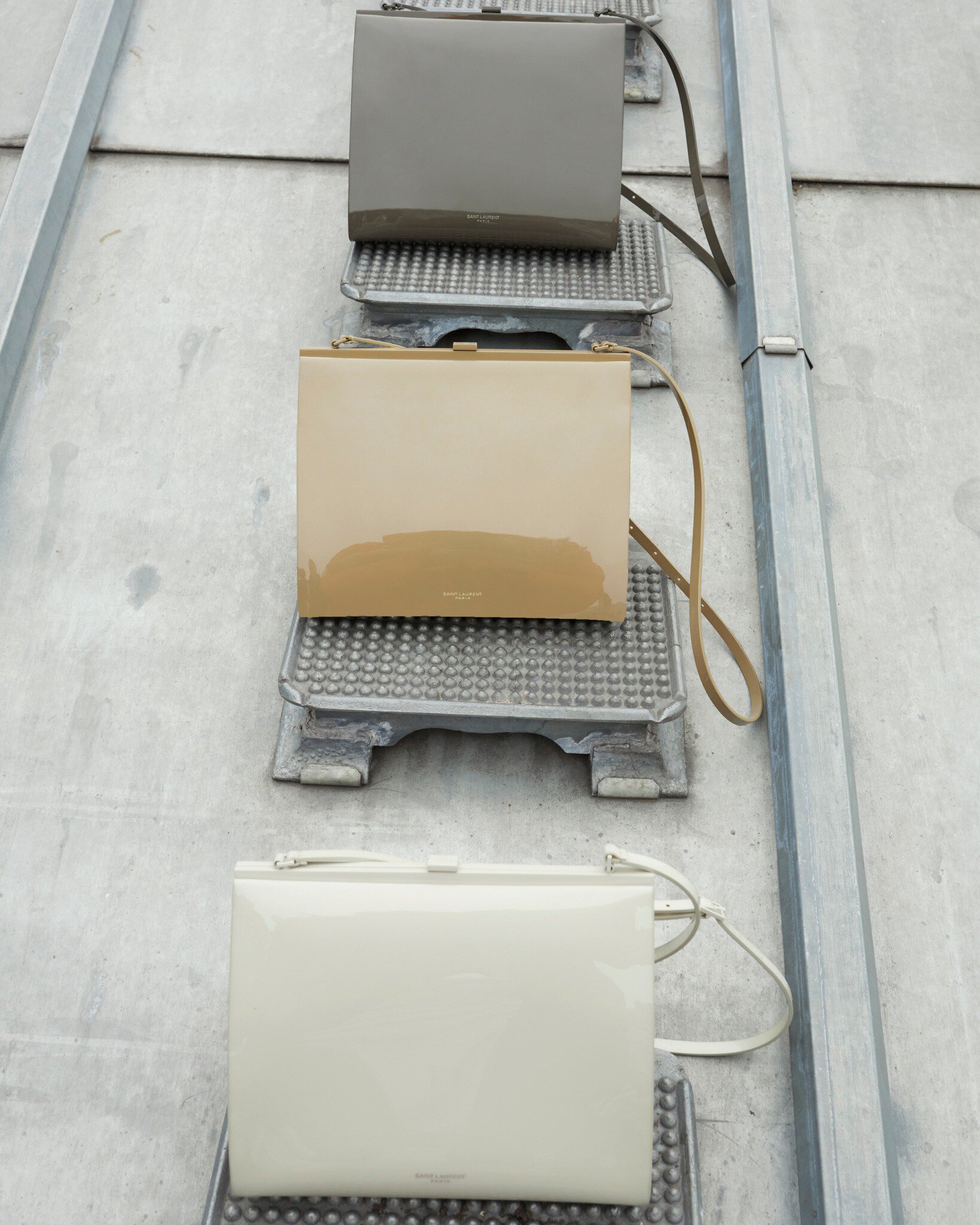 Three glossy, rectangular handbags in graduated neutral tones are staged on metal platforms against a textured, grey, metallic background.