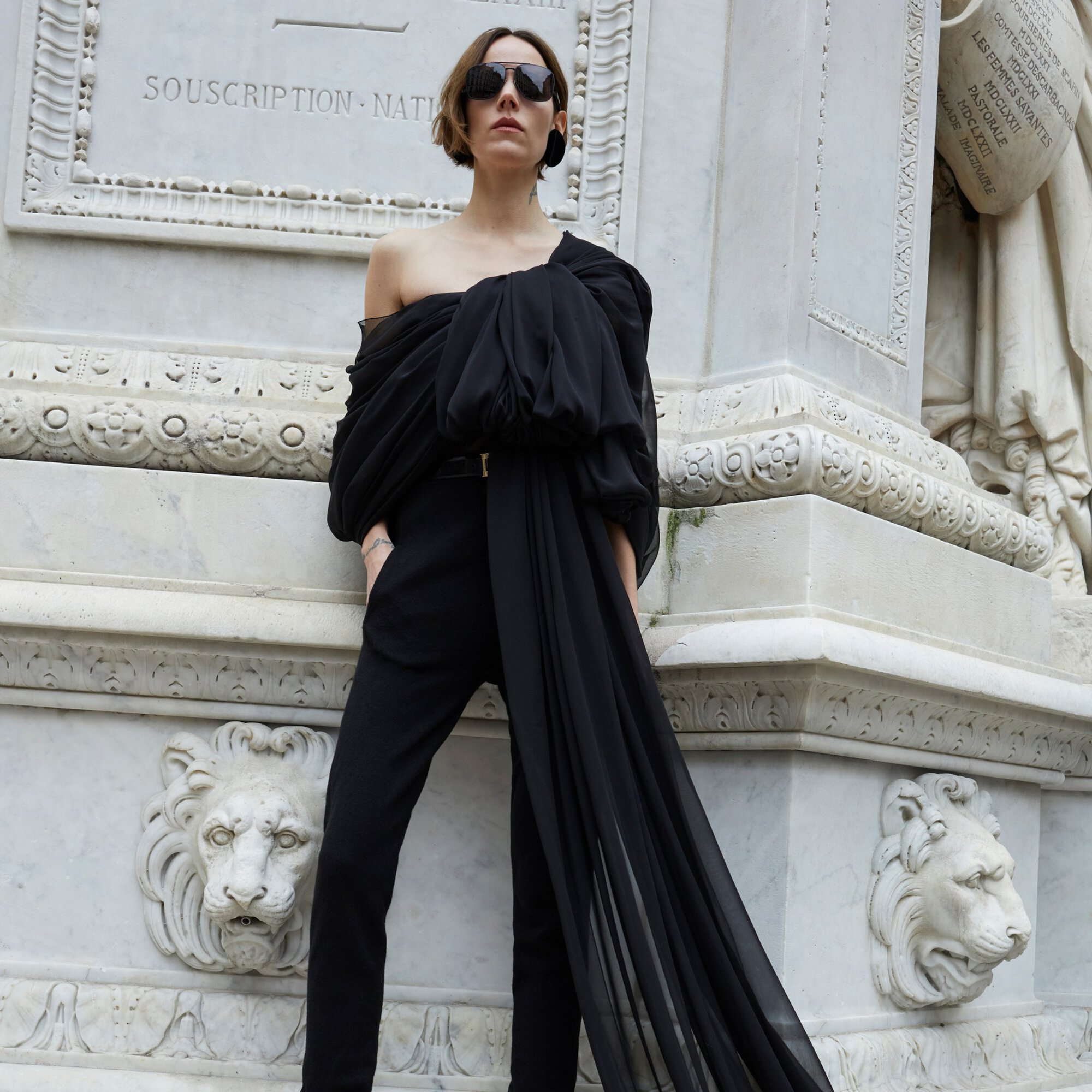 A stylish woman in black, off-shoulder blouse and trousers poses outdoors against a marble monument backdrop, completing the look with sunglasses.
