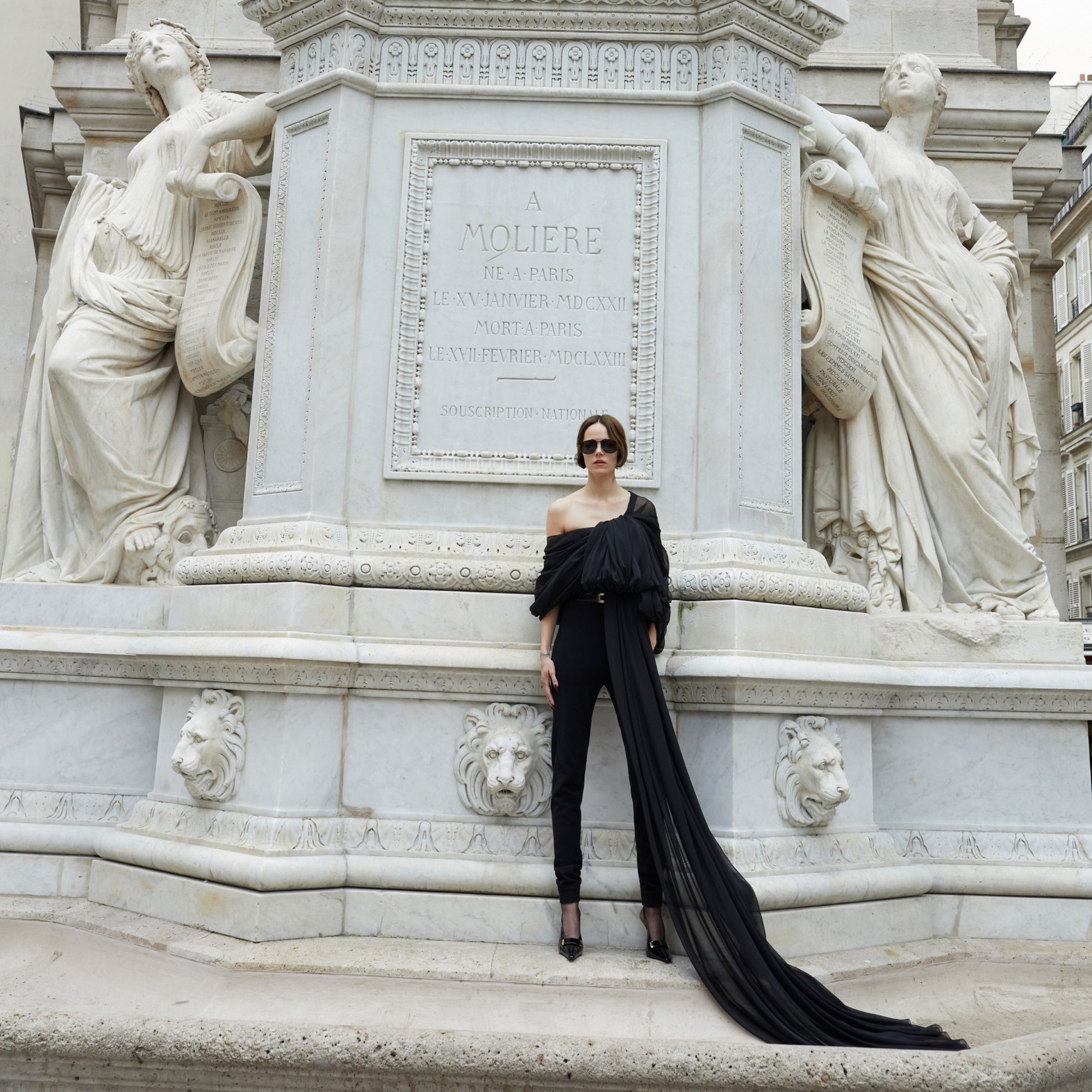 Elegant woman in black jumpsuit poses near monument with statues. Parisian urban setting. Fashionable editorial image.