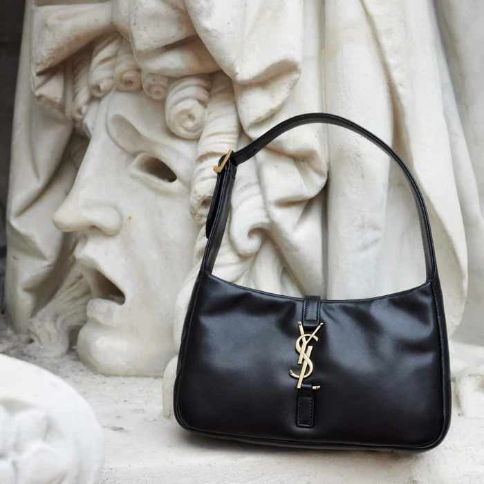 Black leather Saint Laurent shoulder bag with gold monogram hardware resting in front of a white classical marble sculpture.
