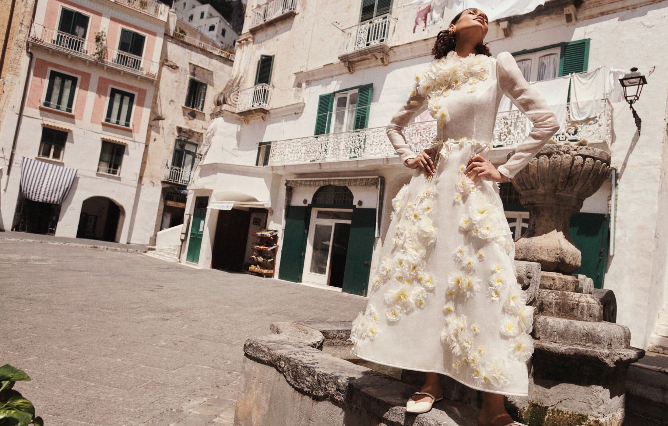 Woman in a floral gown stands in a sunny piazza of a European village with white buildings and stone pavement on a bright day.