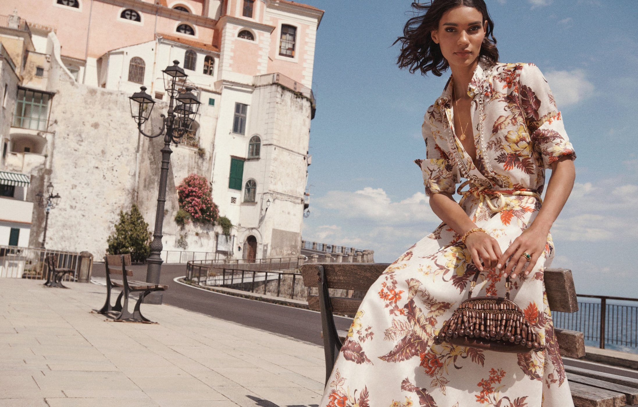 Woman in floral dress sits on stone bench in an Italian town, featuring white buildings, a black lamppost, and bright blue skies.