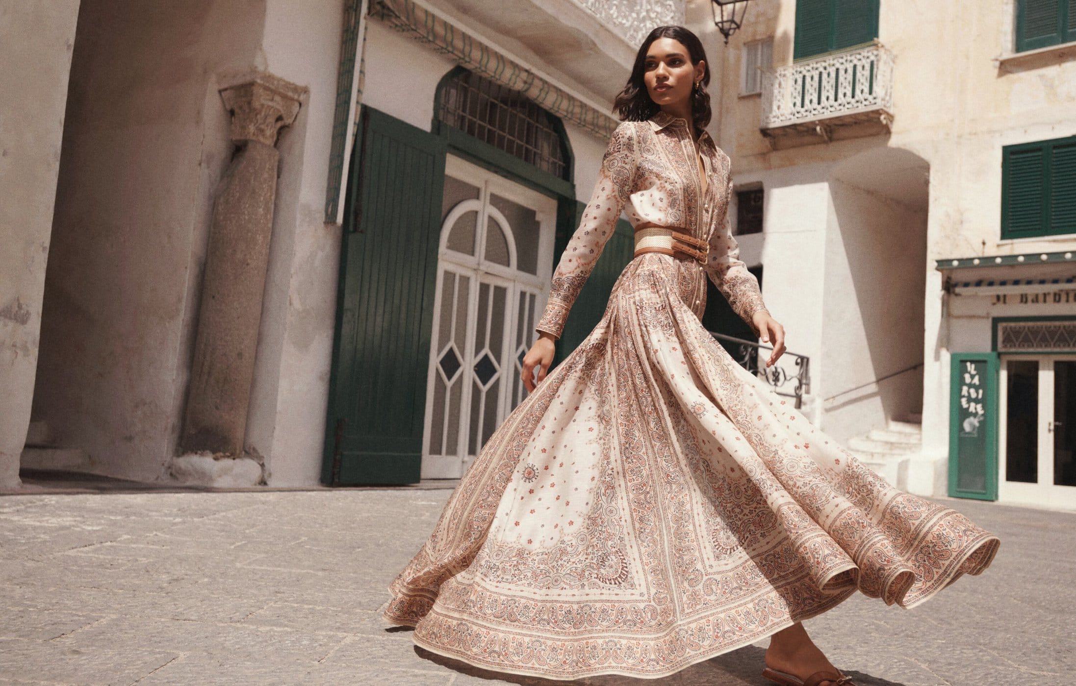 A woman models a long-sleeved, floor-length patterned dress with a wide belt in an outdoor urban setting with neutral-toned architecture.
