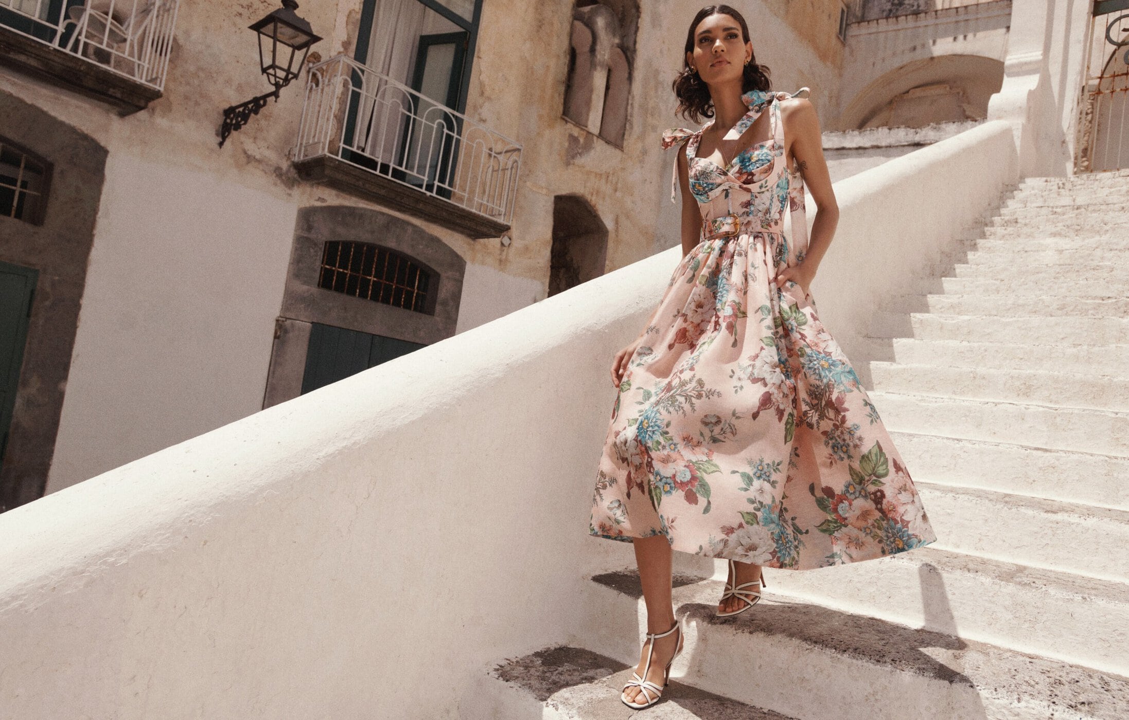Woman in floral dress walks down outdoor stairs in Italian town; sunlight, shadow, warm tones, architectural background.