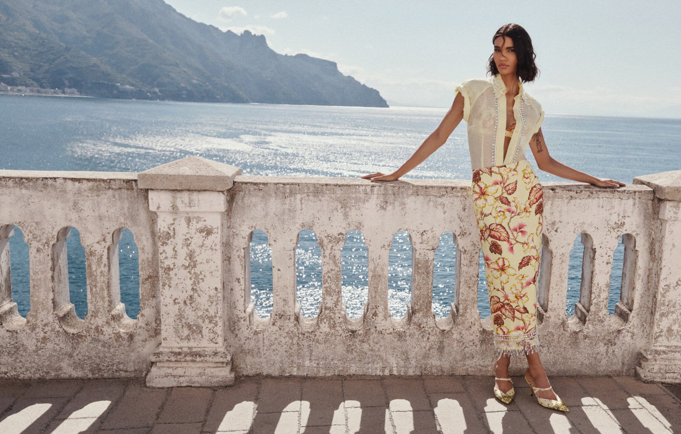 A woman poses on a weathered balcony overlooking the sea, wearing a sheer blouse and floral pencil skirt, exuding elegance in natural light.