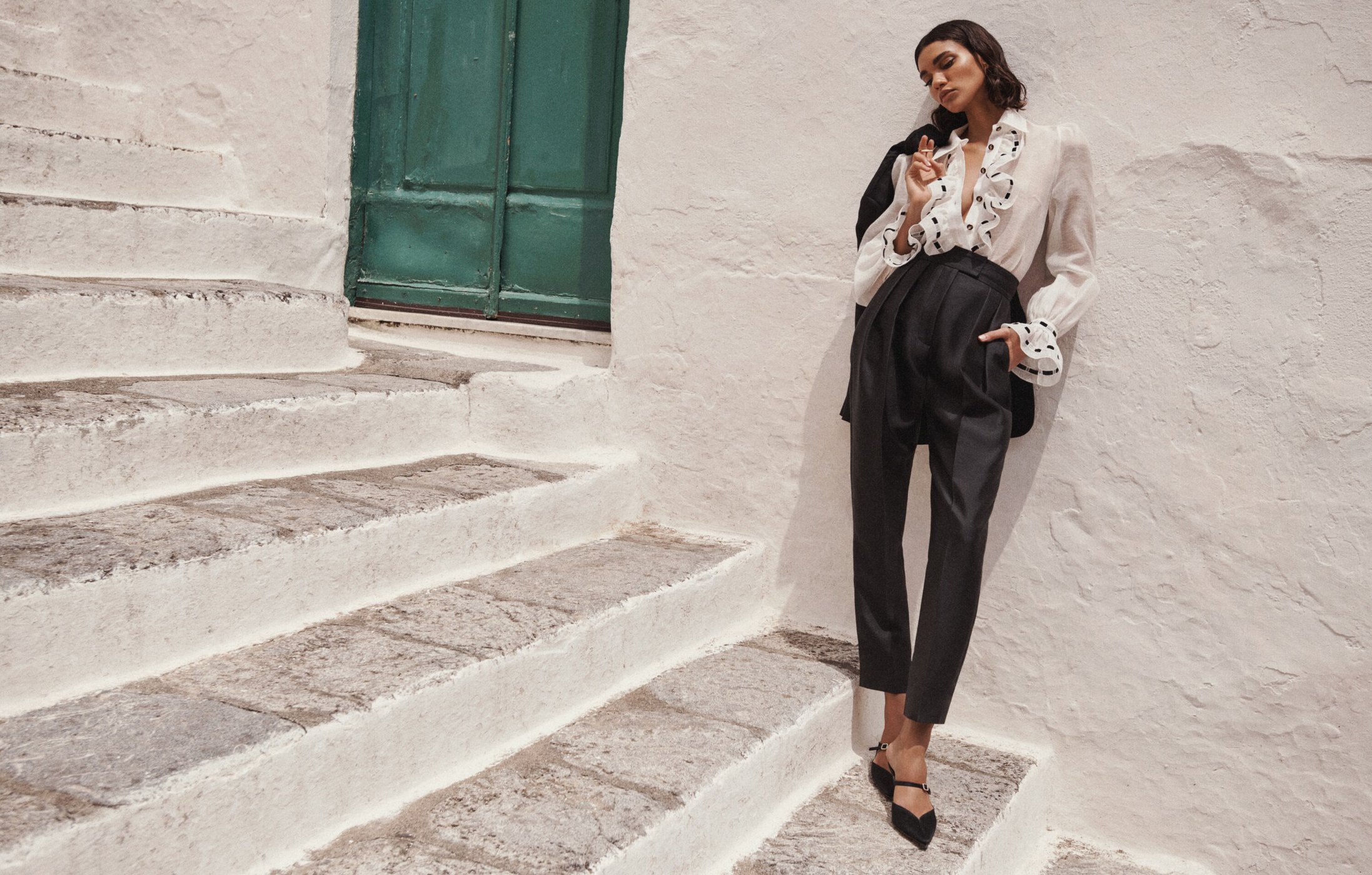 Fashion shot of a slender adult woman posing in a white ruffled blouse and black pants, leaning against a white stucco wall next to stairs.