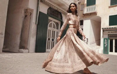 Model Barbara Valente spins in an ornate cream maxi dress with a wide brown belt in a sunlit Italian public square.