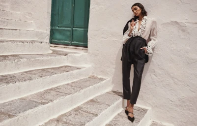 Model Barbara Valente wearing high-waisted black trousers and a white ruffled blouse on stone steps against a white stucco wall.