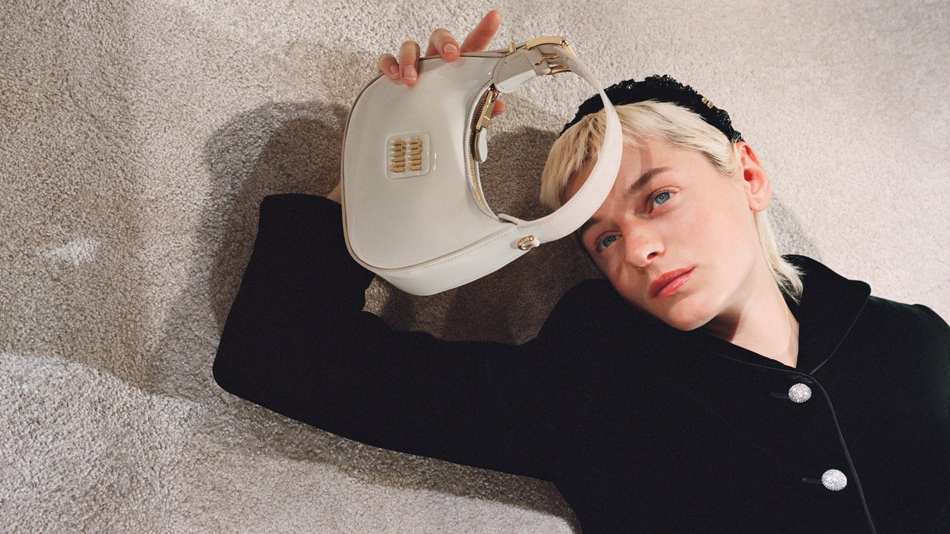 Fashion portrait of a fair-skinned young adult with a platinum blonde hair, holding a white Miu Miu handbag on a carpet background.