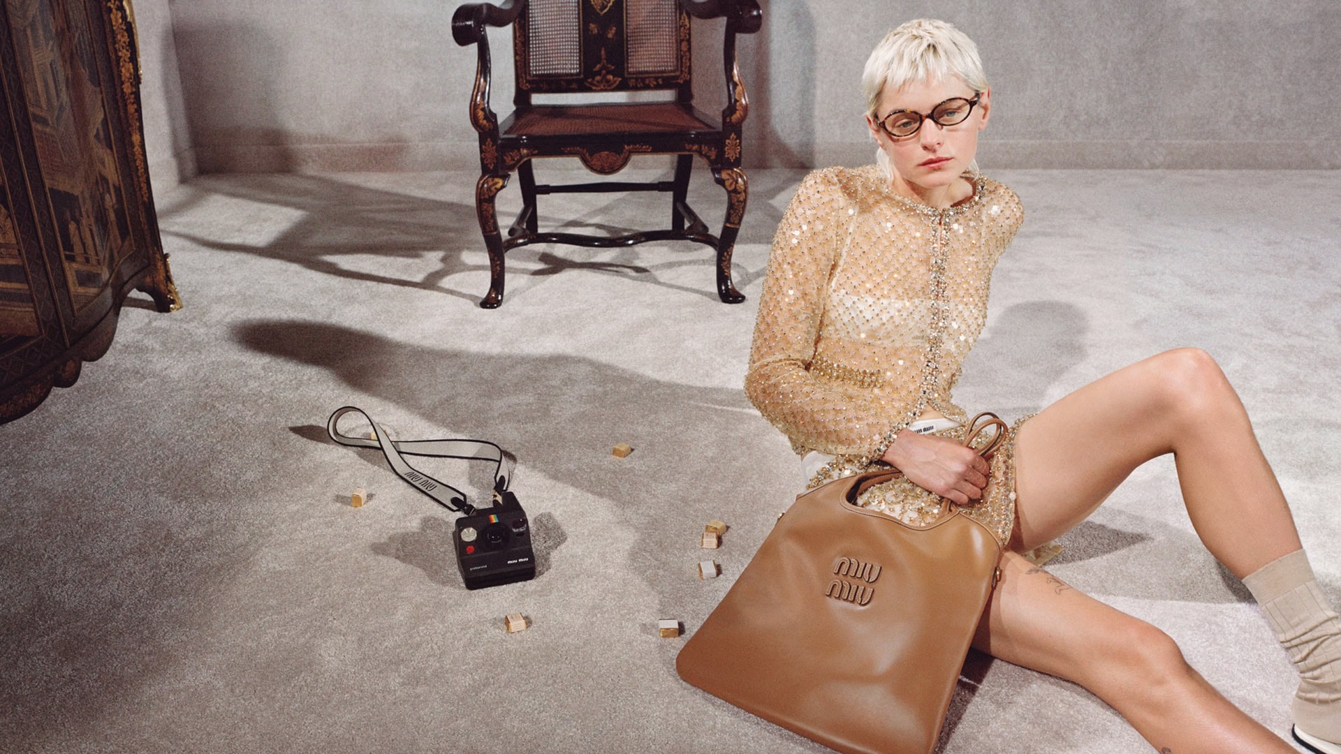 Fashion shot of a woman seated on a carpeted floor in front of vintage furniture with a Miu Miu handbag and instant camera.