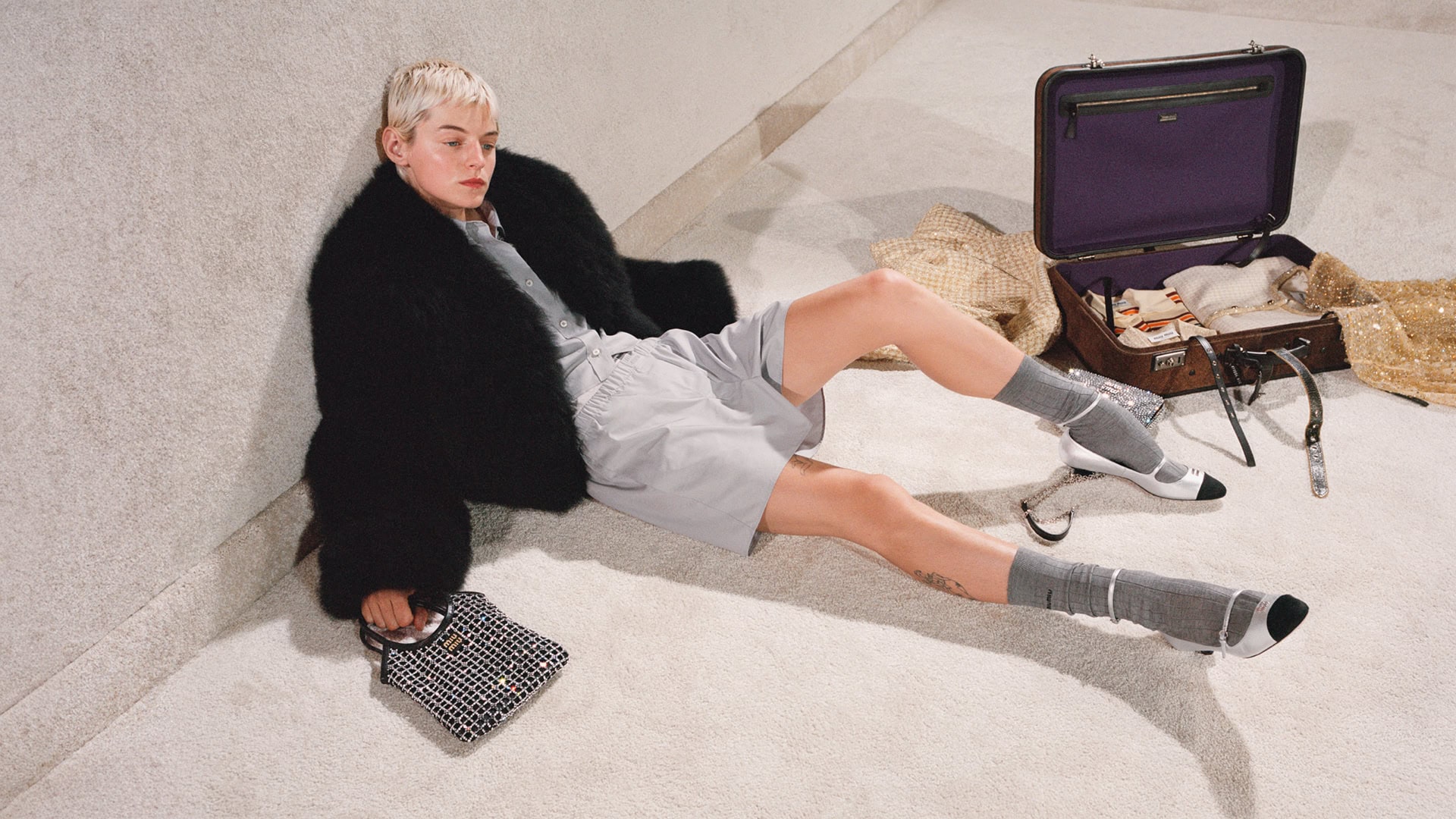 Fashionable woman with short blonde hair sitting on floor in black fur coat, gray shorts, and heels, with handbag and suitcase.