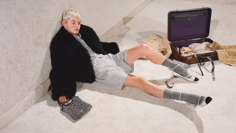 Fashionable woman with short blonde hair sitting on floor in black fur coat, gray shorts, and heels, with handbag and suitcase.