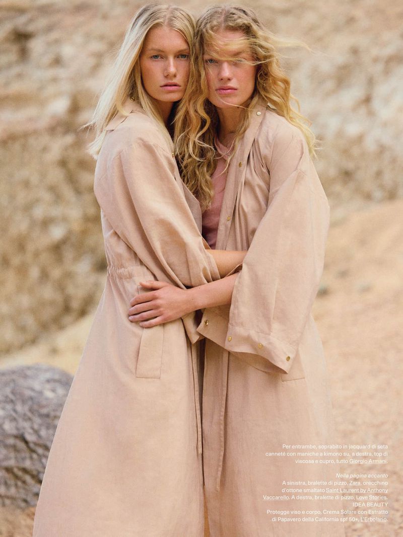 Two blonde women in tan trench coats embrace in a sunlit desert landscape. Fashion editorial, neutral tones, outdoor setting, long wavy hair.