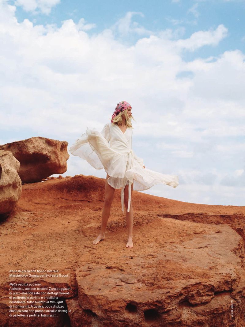 Fashion shot of a woman in a flowing white dress and headscarf posing on red rocks against a blue sky with clouds, outdoors.