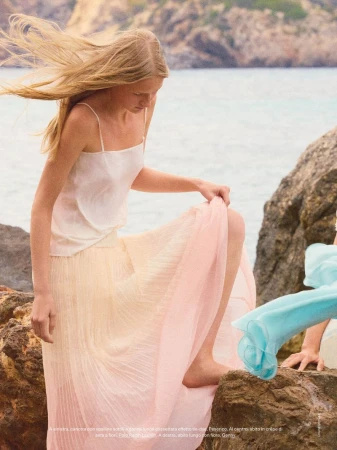 A blonde woman in a flowing skirt poses on rocks by the sea, with windblown hair, creating a serene and fashionable oceanside scene.