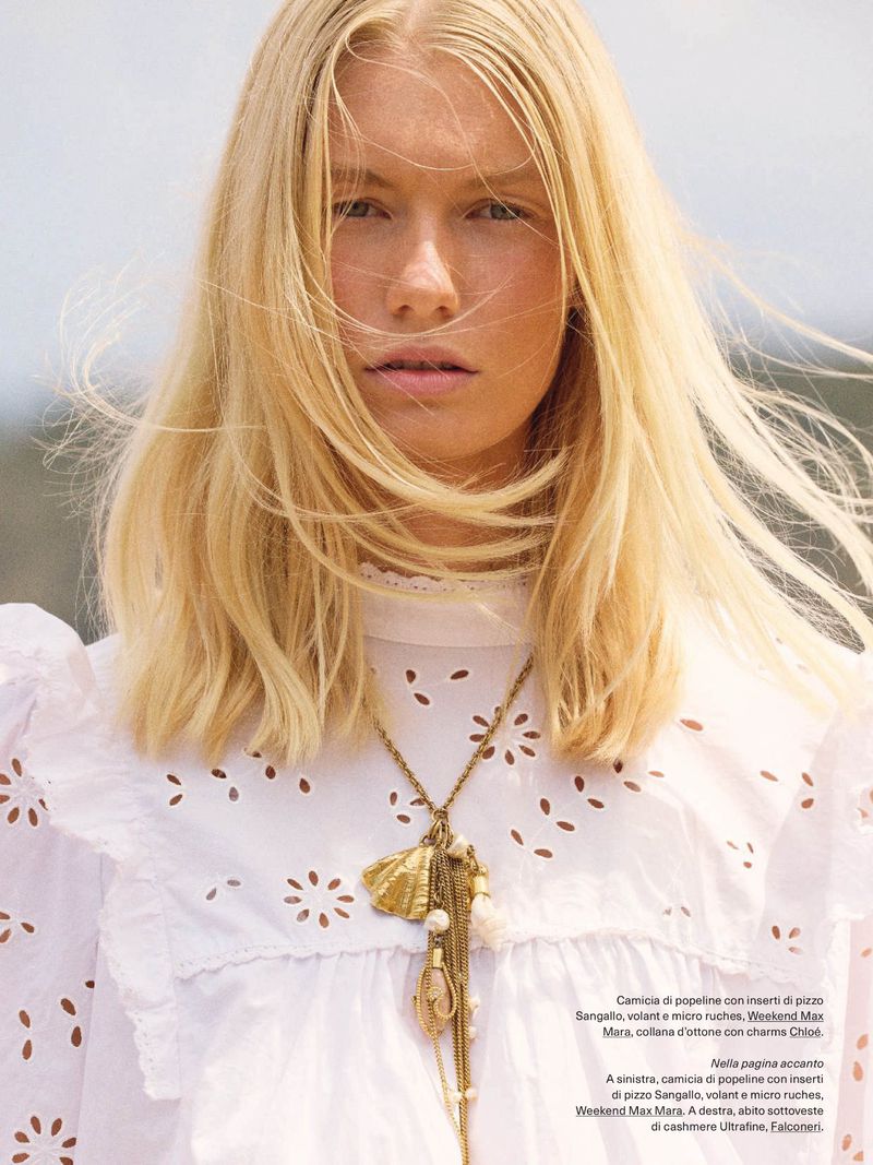 Close-up fashion shot of a blonde adult woman wearing a white eyelet dress and shell necklace, with a neutral outdoor background.
