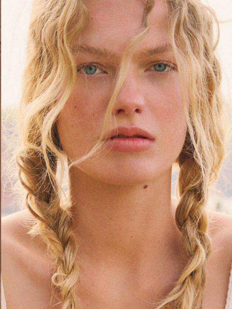 Close-up portrait of a woman with fair skin, blue eyes, and blonde curly hair styled in braids, looking directly at the camera.