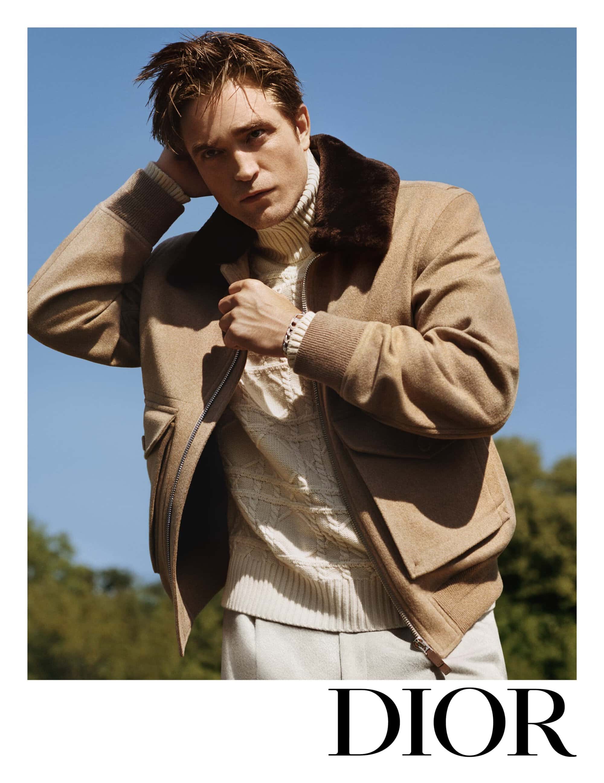 A man in a fur-collared jacket poses outdoors against a clear blue sky. He wears a cable-knit sweater and light-colored pants.