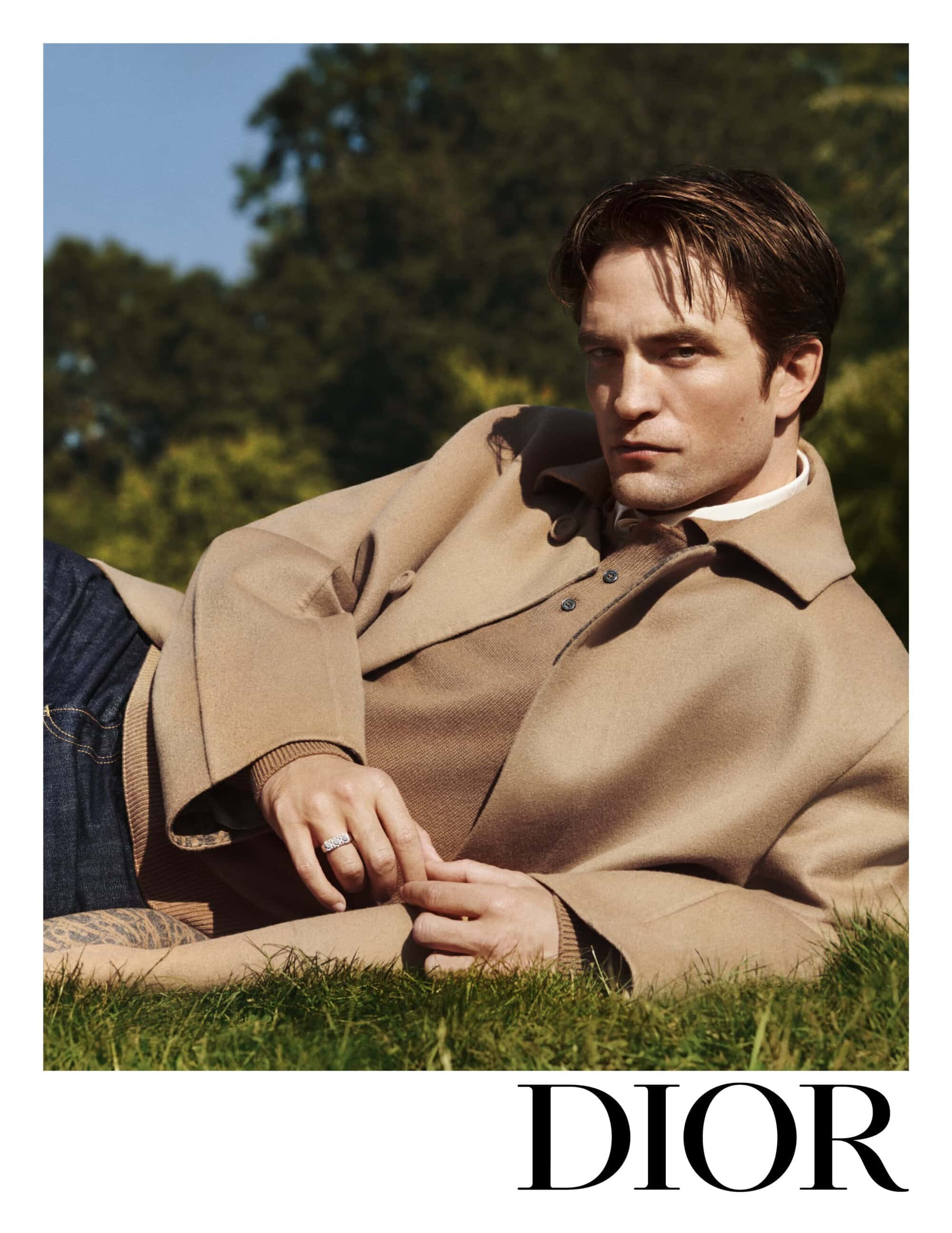 Robert Pattinson lies on a grassy lawn, wearing a beige coat and looking at the camera in a Dior advertisement, surrounded by nature.