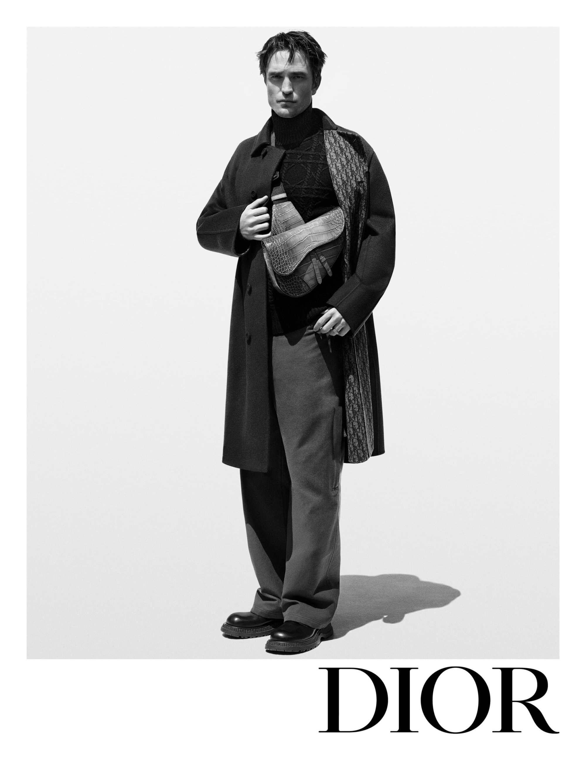 Black and white portrait of male fashion model Robert Pattinson in overcoat holding textured saddle bag in a bright photo studio.