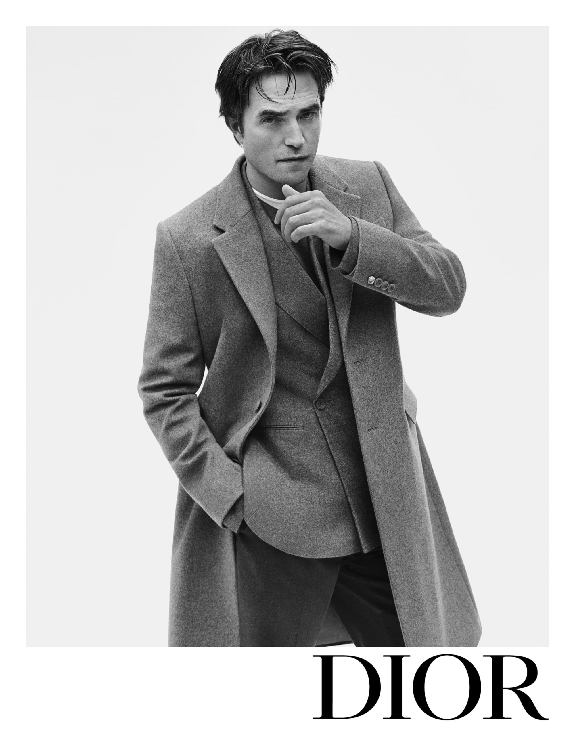 Black-and-white portrait of adult male, Robert Pattinson, wearing a suit and long coat, posing in a photo studio with bright, diffused lighting.