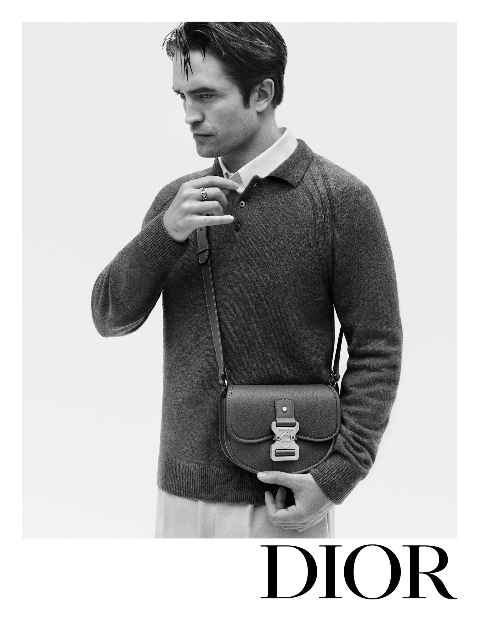 Black and white portrait of Robert Pattinson wearing a grey sweater, carrying a small saddle bag, with the Dior logo at the bottom.