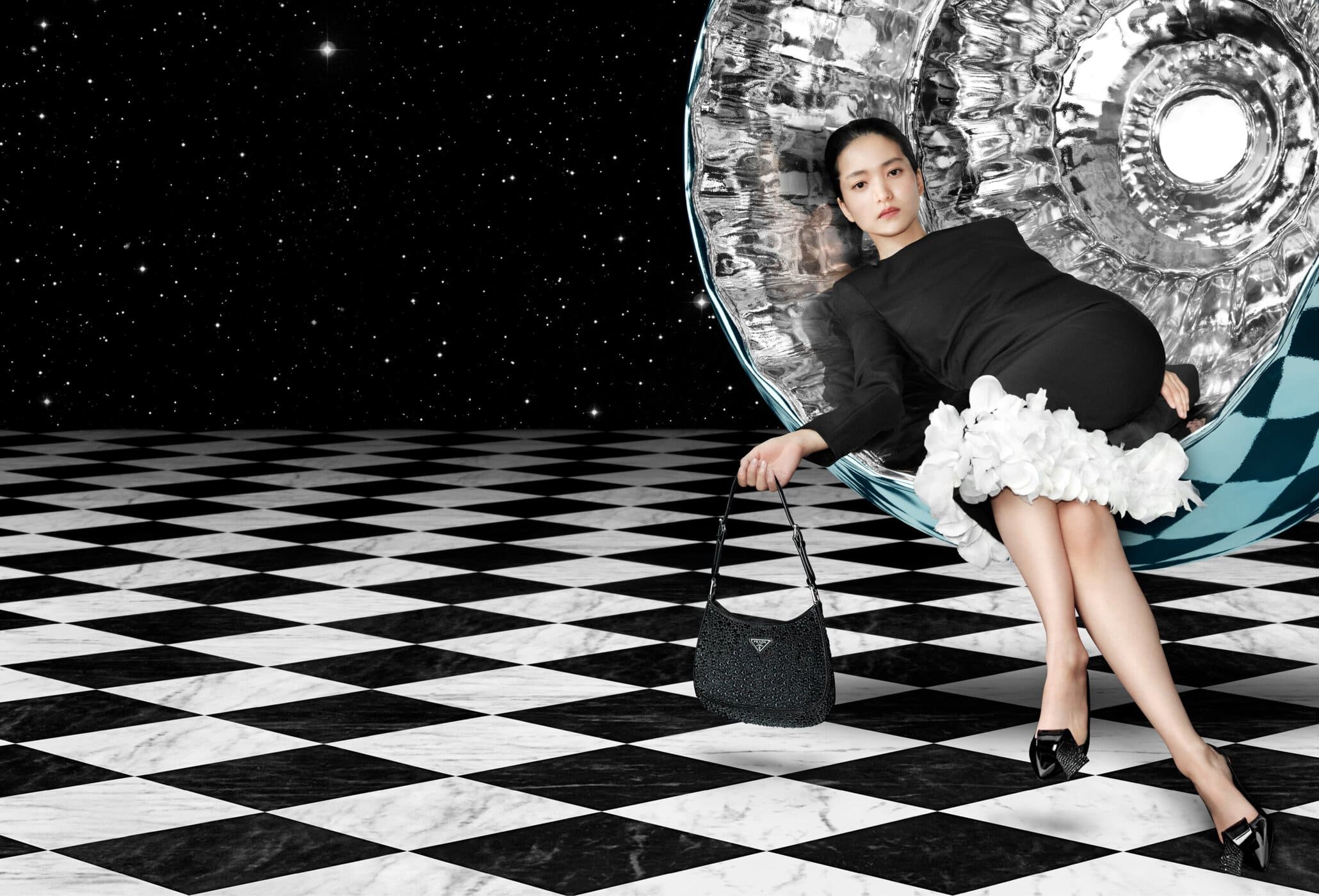 A woman in a black dress poses with a black Prada purse in a surreal, space-themed studio setting featuring a checkered floor.