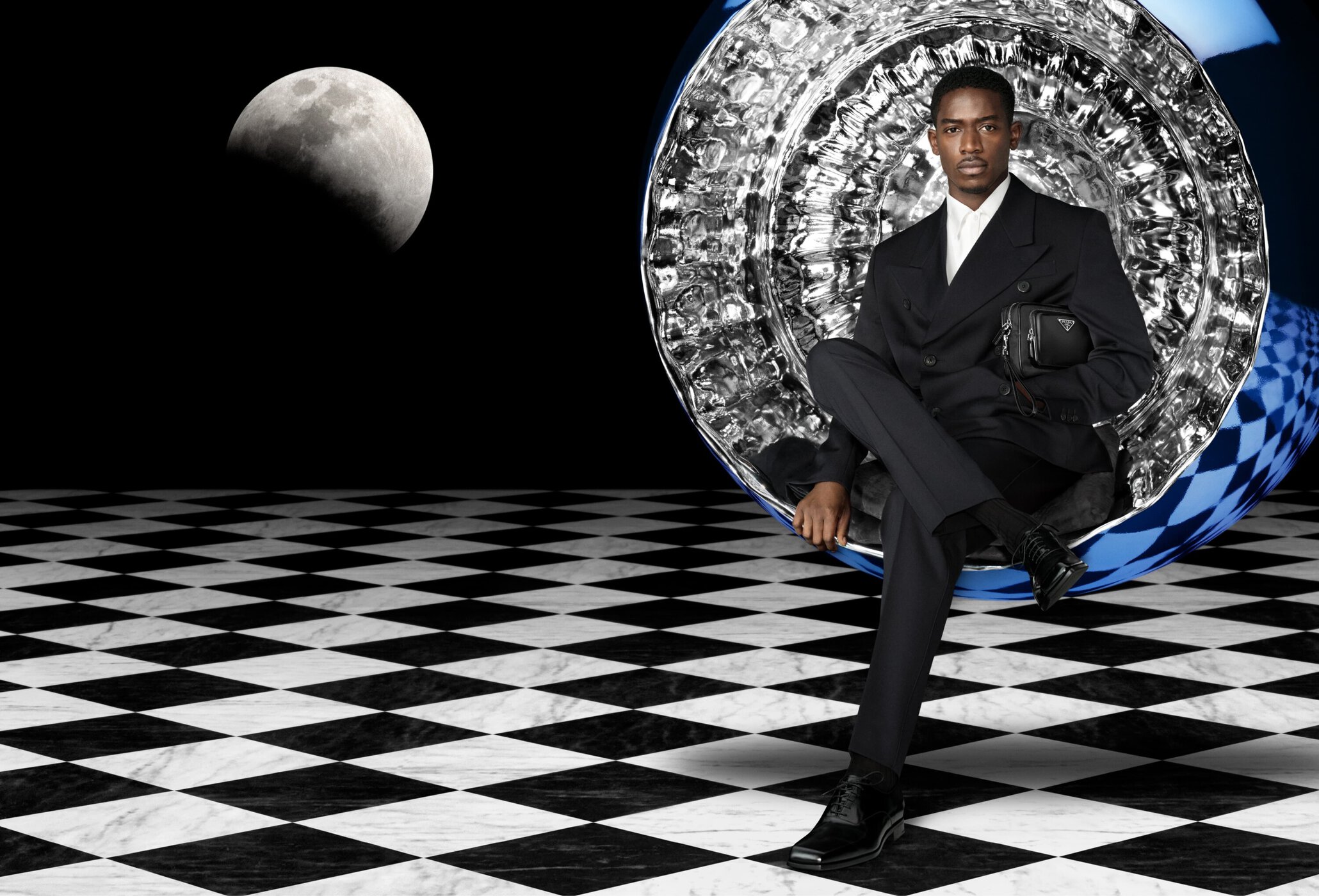 Man in black suit poses in futuristic chair in front of moon on a black and white checkered floor, holding a black Prada bag.