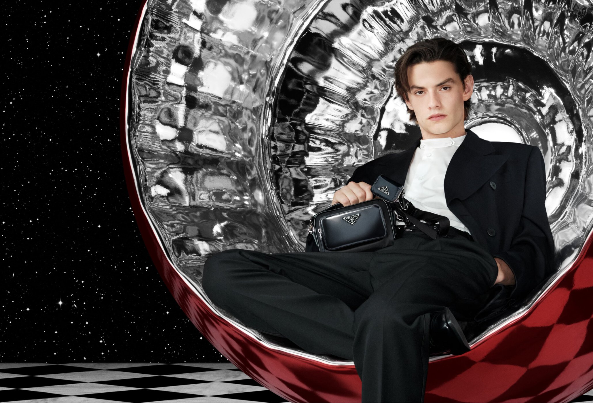 Man in a black suit sits in a reflective silver chair against a star field backdrop, holding a black Prada bag, looking at the camera.