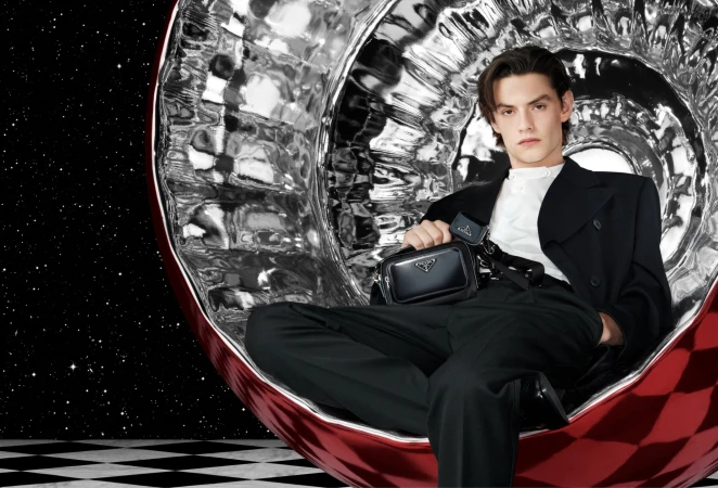 Man in a black suit sits in a reflective silver chair against a star field backdrop, holding a black Prada bag, looking at the camera.