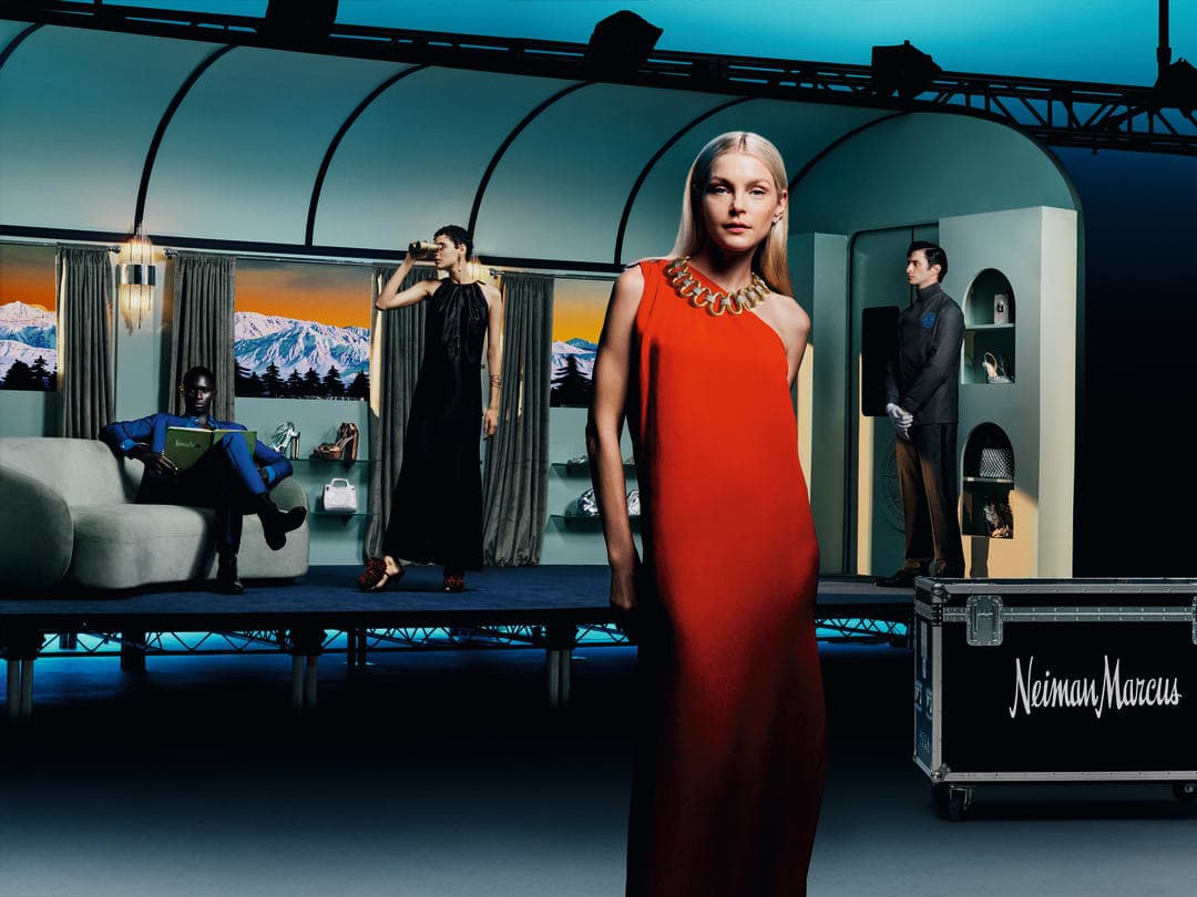 Fashion editorial shot in an indoor studio setting featuring a blonde female model wearing a red dress and gold necklace, with other models on a platform.