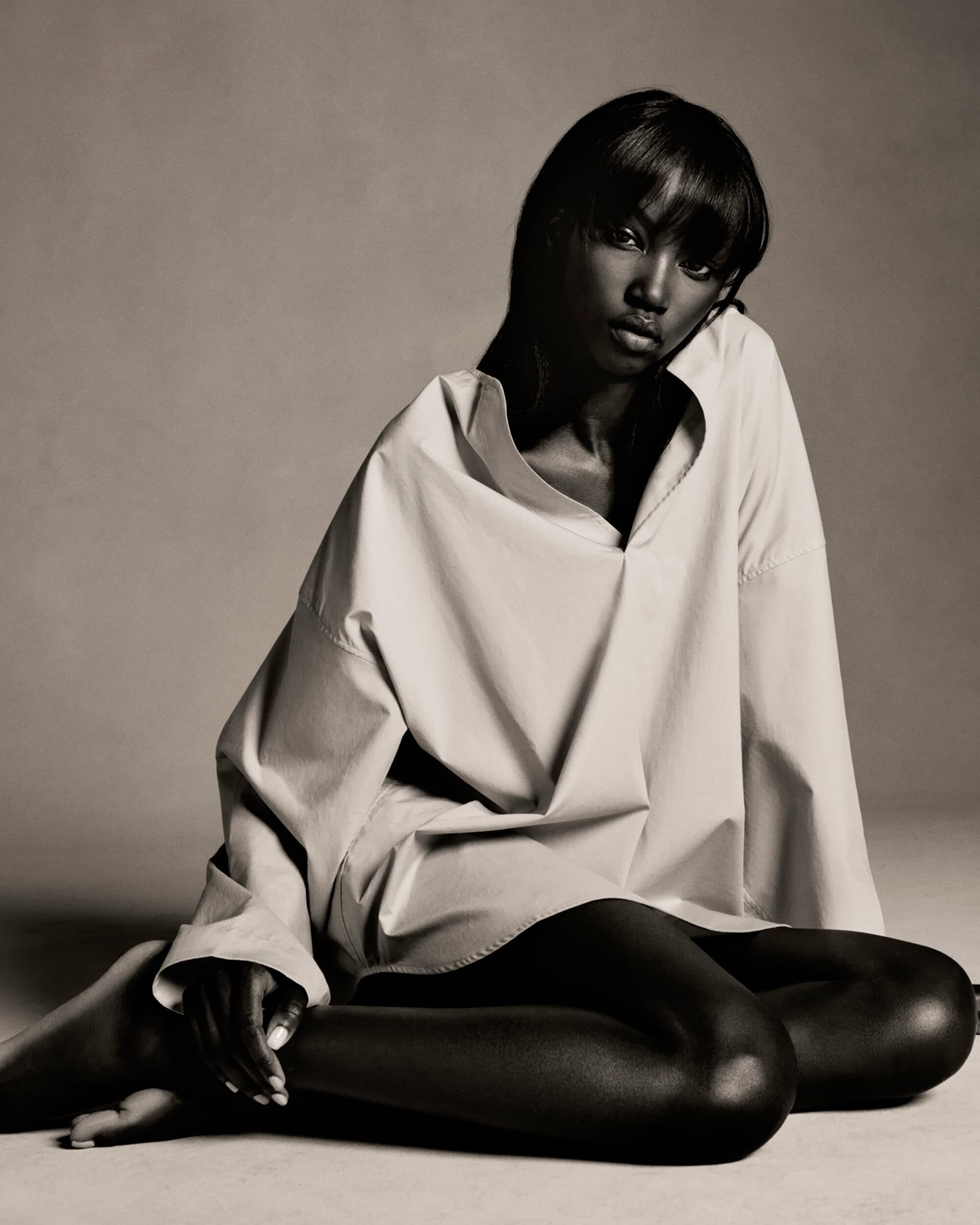 Monochrome fashion portrait of a dark-skinned woman in an oversized shirt, sitting in a photo studio with a serious expression.