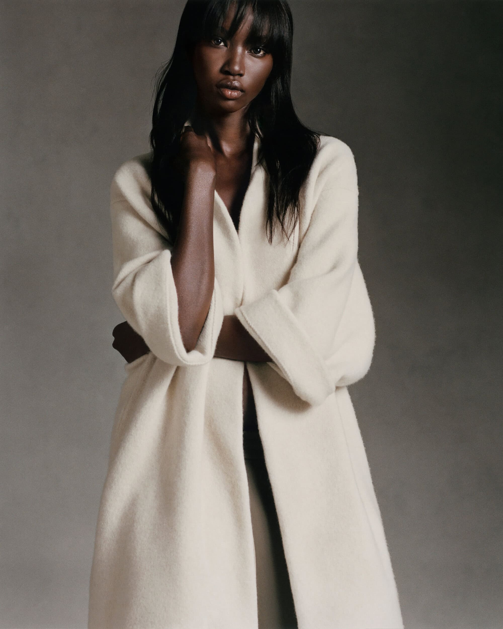 Dark-skinned woman poses in a cream-colored wool coat against a neutral background, looking directly at the camera with a serious expression.
