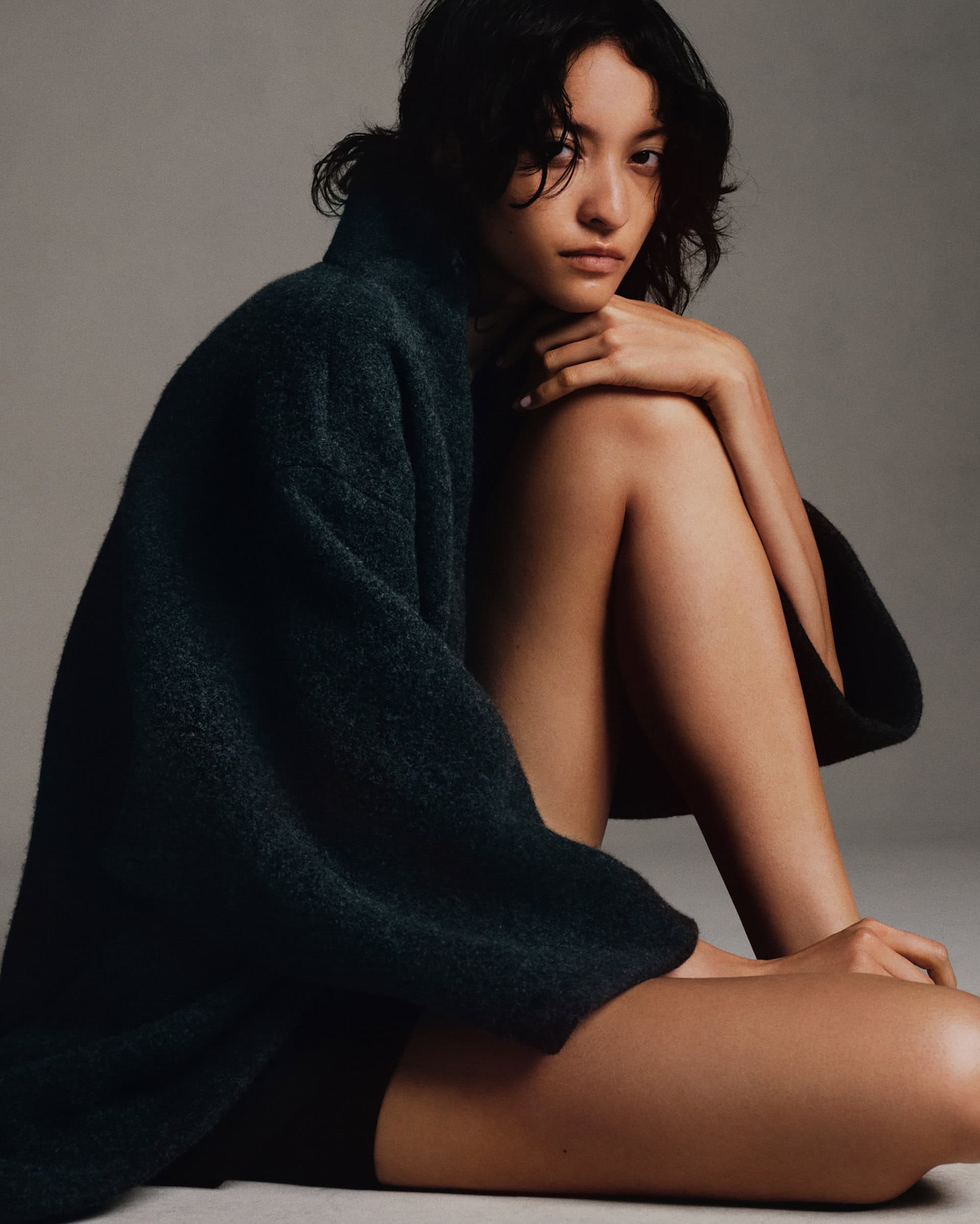 Young woman with dark hair sits in a casual pose wearing a dark green knitted wool cardigan in a photo studio with soft lighting.