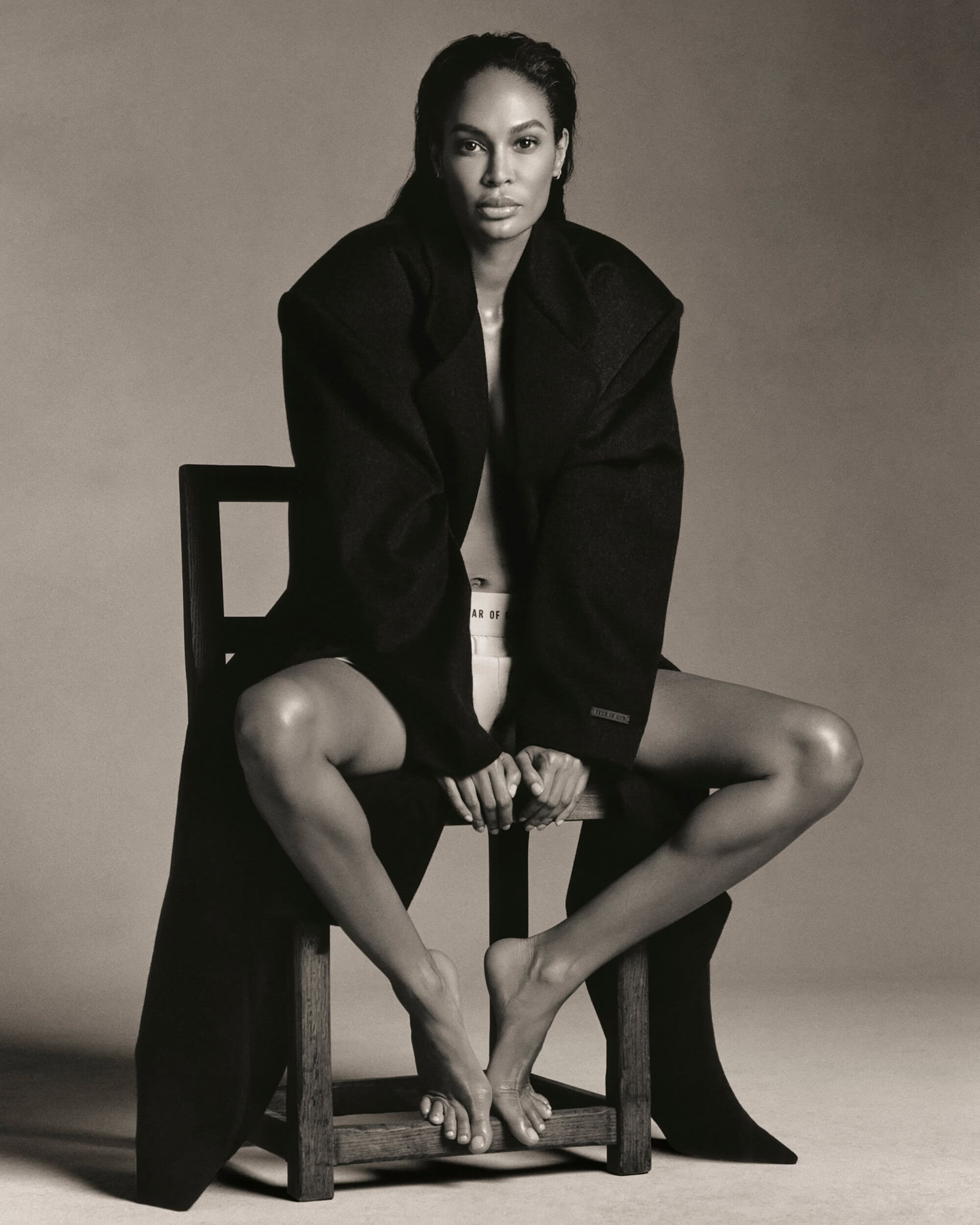 Elegant woman in a black coat strikes a pose while seated on a wooden chair in a sepia studio portrait; fashion and beauty concept.