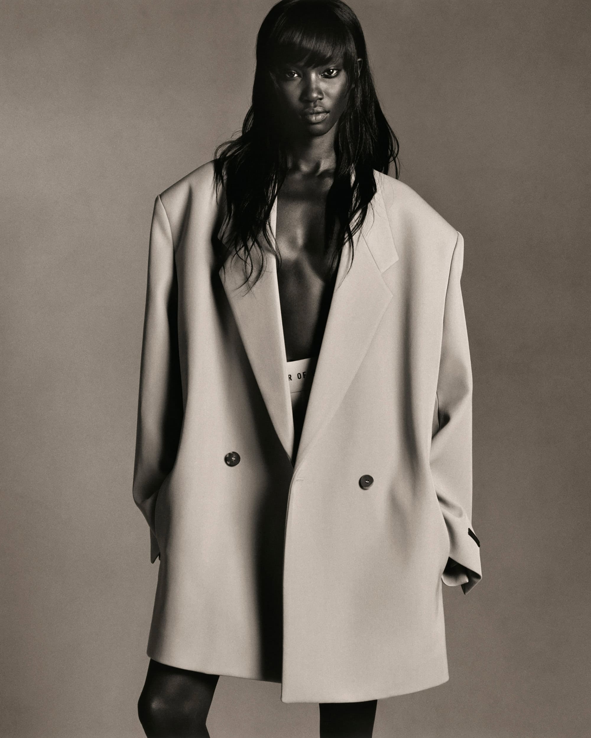 Portrait of an adult Black female model wearing an oversized beige double-breasted blazer looking at the camera in a photo studio setting.